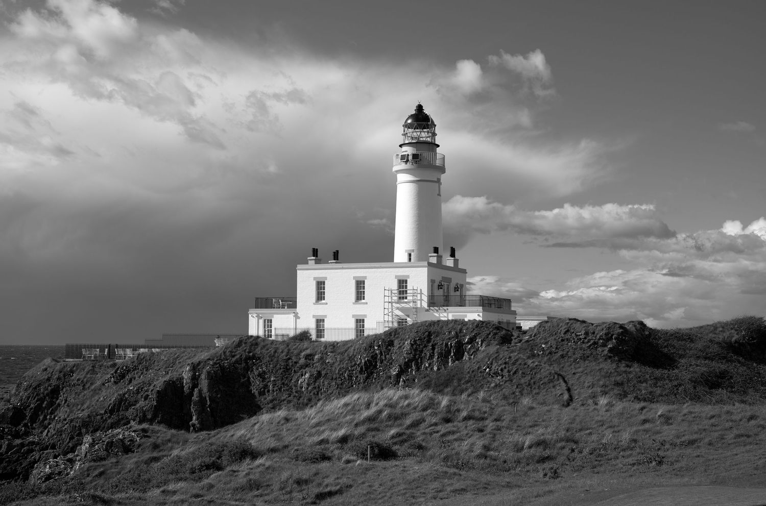 Black and white photo of a white lighthouse on rocky coastal cliffs under dramatic cloudy skies.
