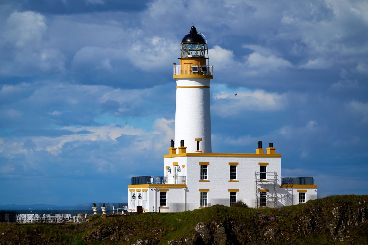 White lighthouse with yellow accents on rocky Scottish coastline under dramatic blue cloudy sky.