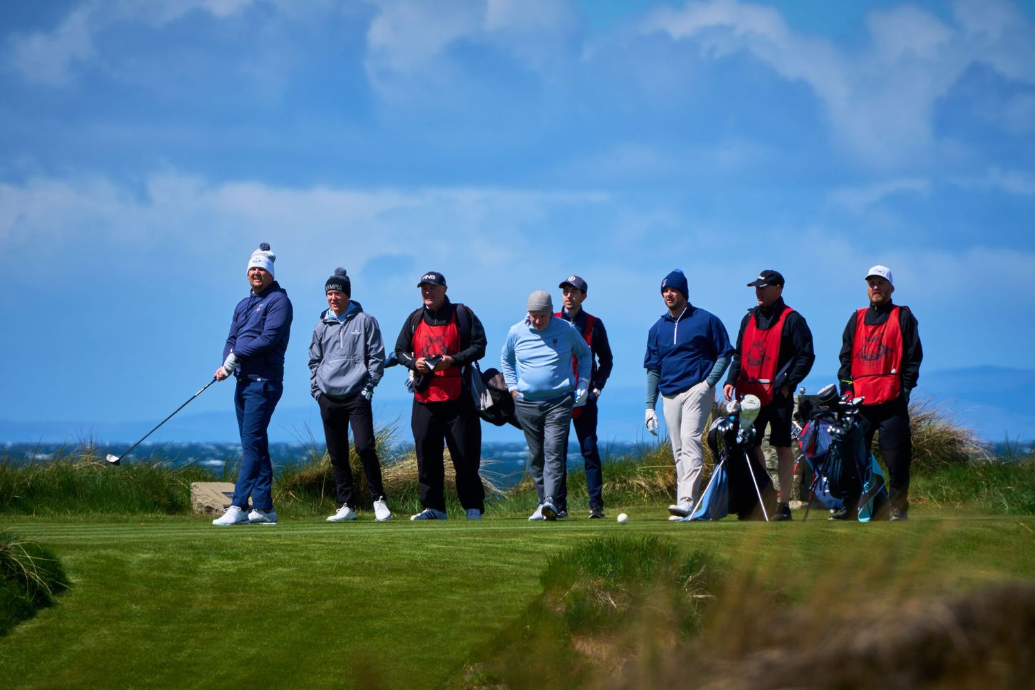 Group of seven golfers and caddies standing on a coastal links golf course under a blue sky.