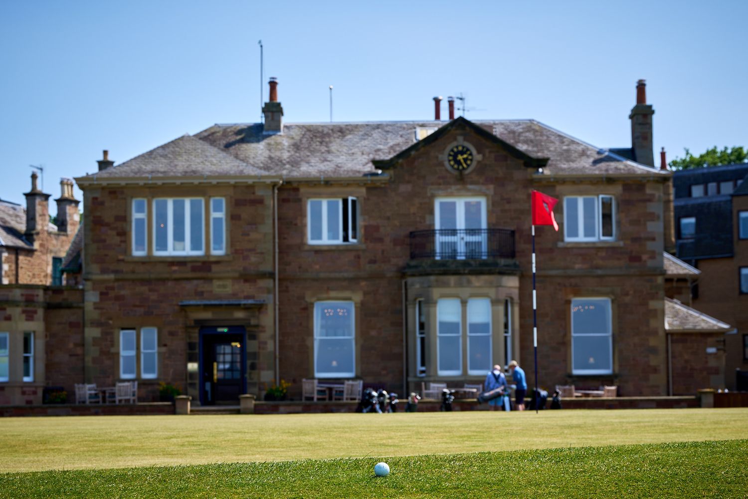 Golfer putting on a green in front of a historic stone clubhouse with a red flag on a sunny day.