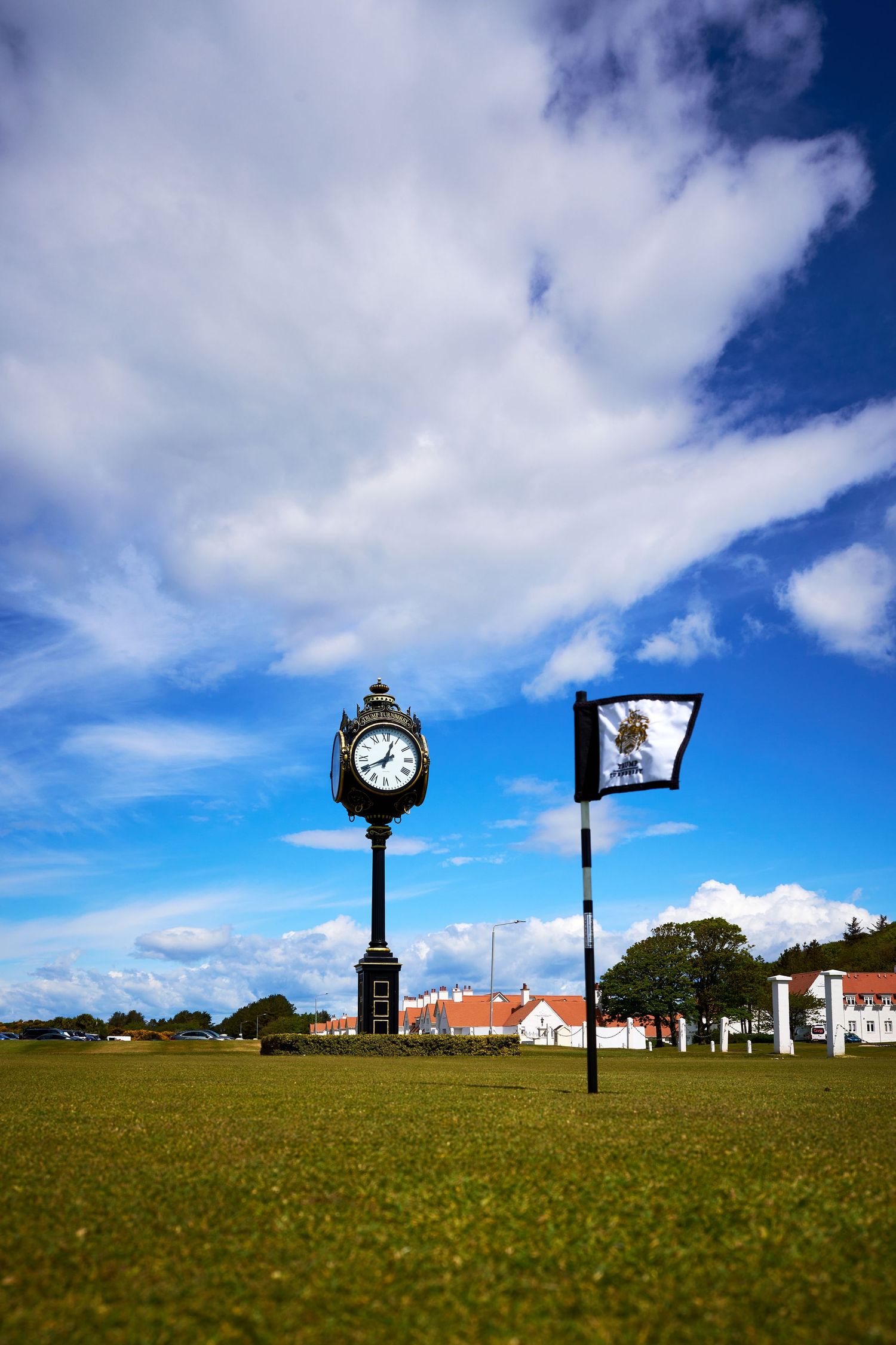 Ornate clock post and flag on lush green golf course under dramatic blue sky with white clouds.