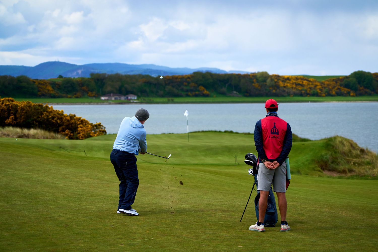 Golfer taking a shot on a scenic coastal golf course with a caddie, water and mountains in the background.