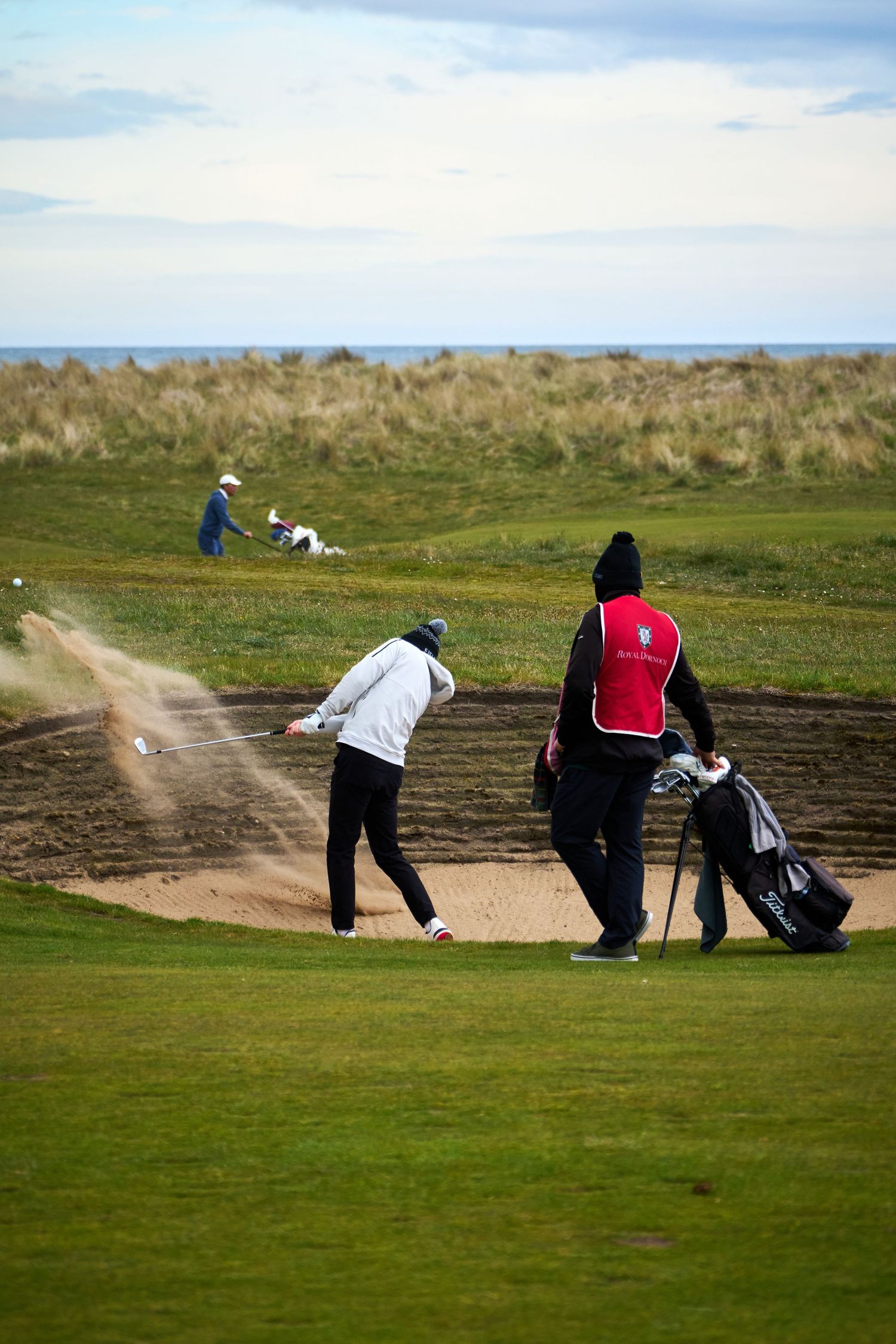 Golfer hitting a bunker shot on a links course with caddie nearby and ocean views in the background.