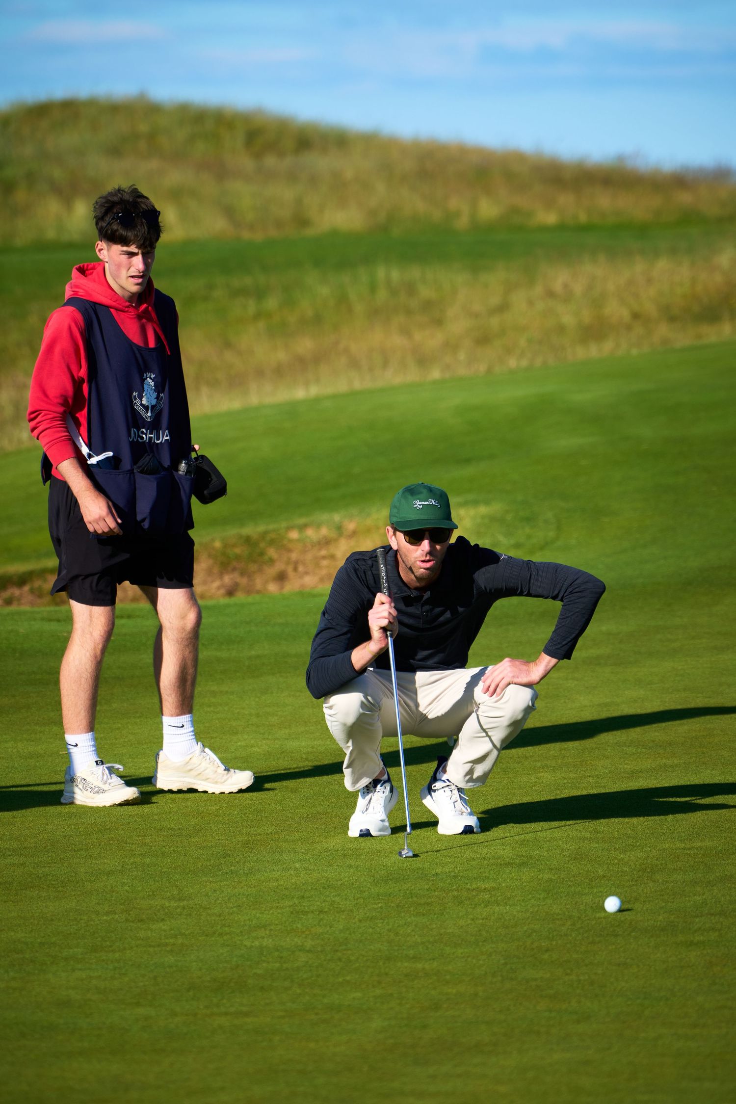 Golfer crouching to read a putt on a green while a caddie in a red hoodie observes on a sunny golf course.