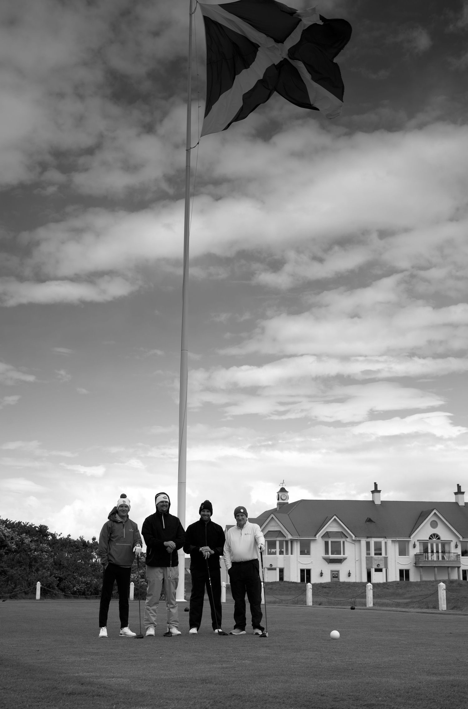 Four golfers posing under a large Scottish Saltire flag on a golf course with a clubhouse behind.