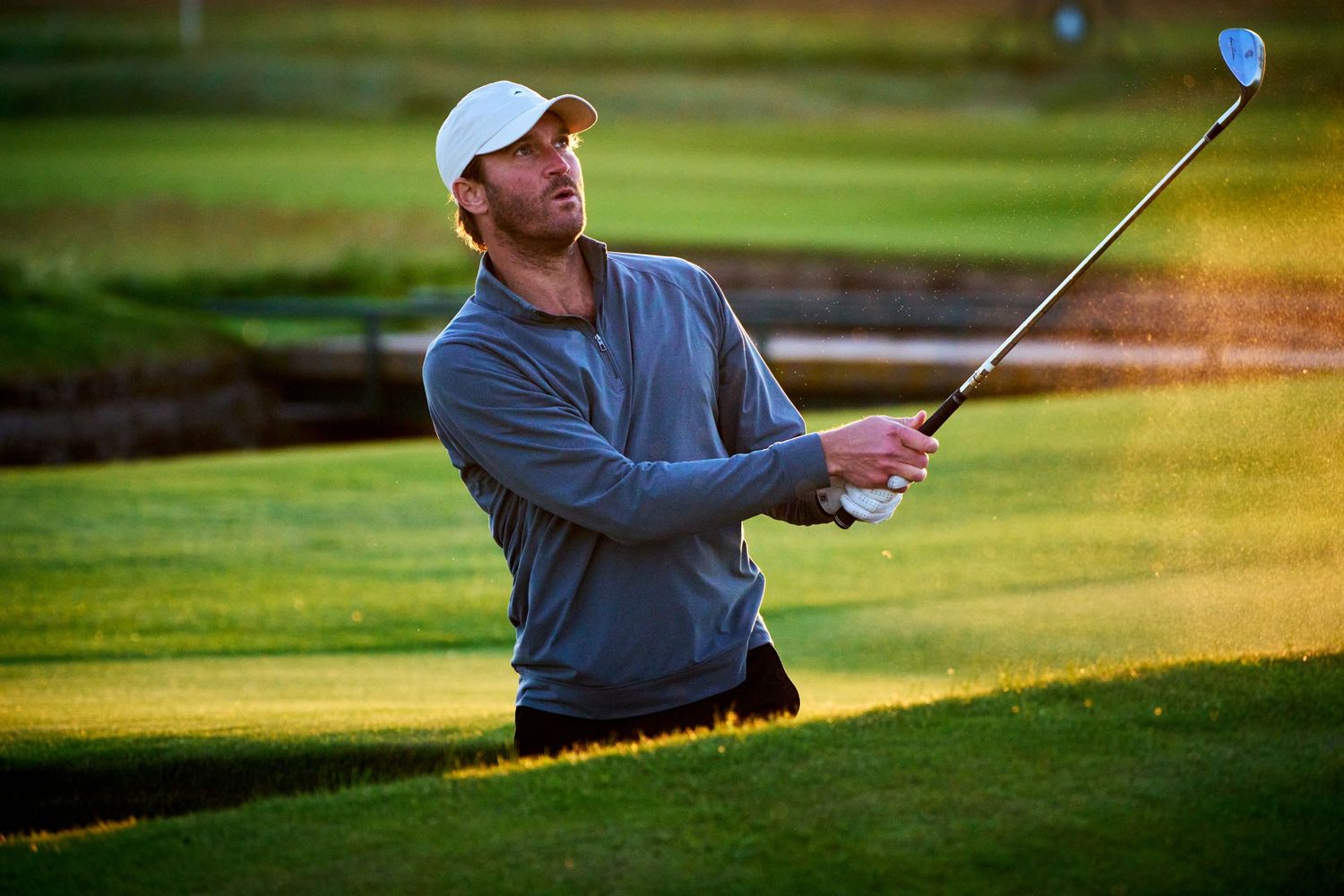 A golfer in a white cap and gray sweater swings a club on a sunlit golf course.