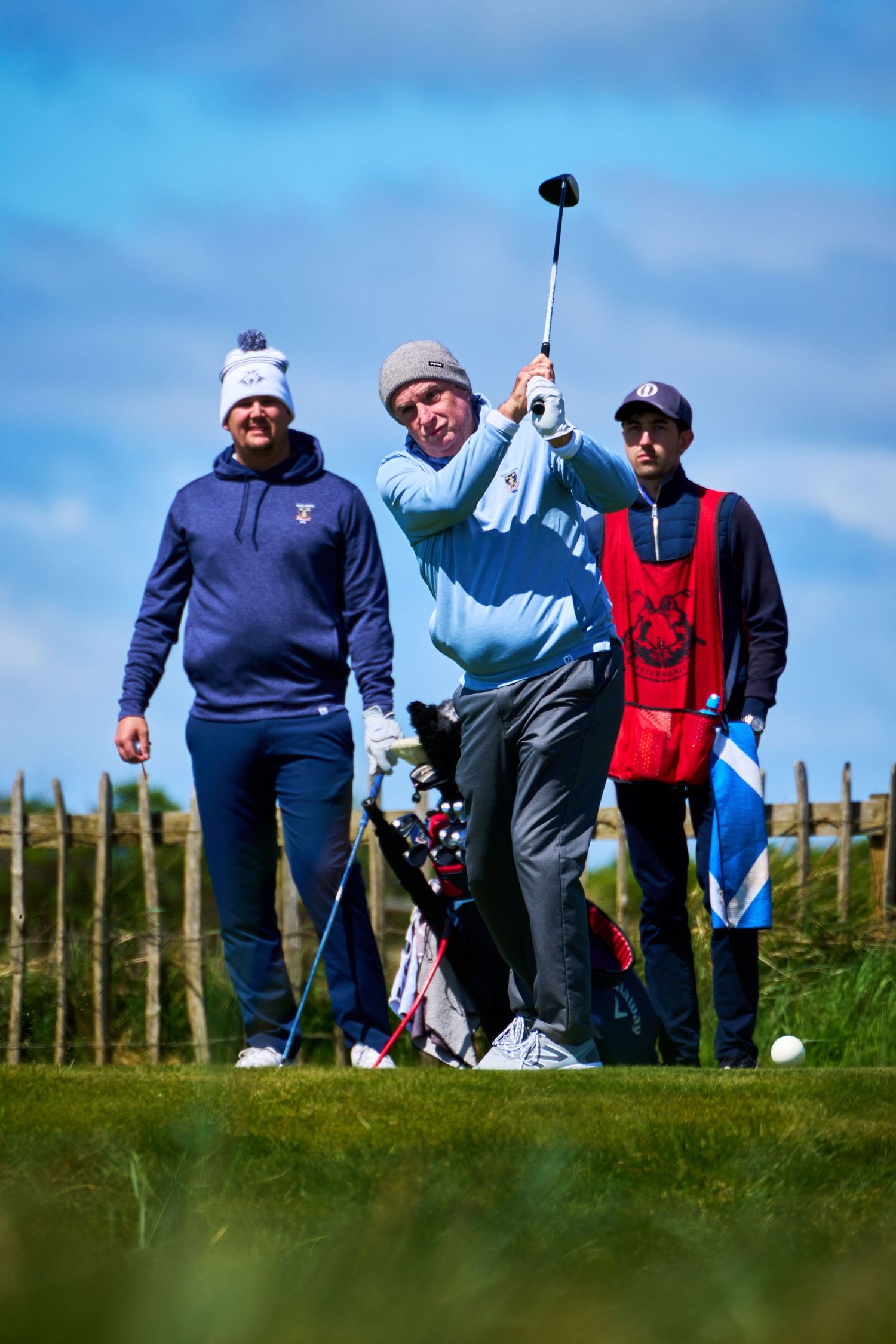 Golfer mid-swing on a links course with caddie and spectator watching under a blue sky.
