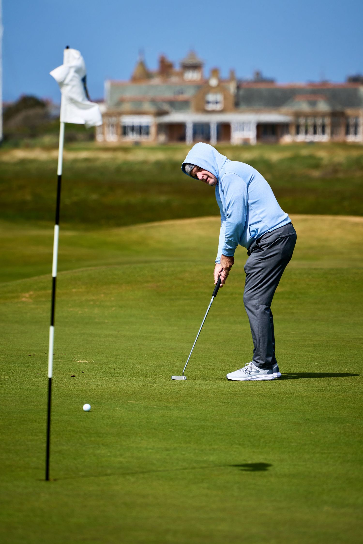 Golfer in blue hoodie putting on a green golf course with a flag and historic clubhouse in the background.