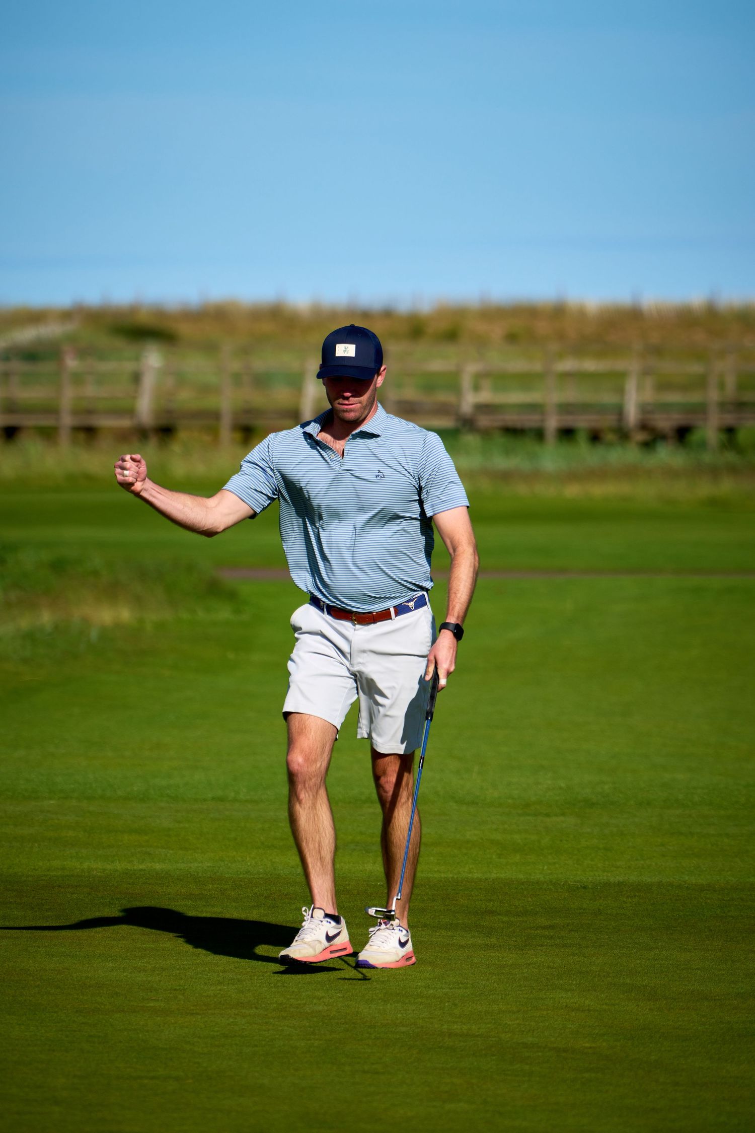 Golfer in blue polo shirt and white shorts walking on a lush green golf course on a sunny day.
