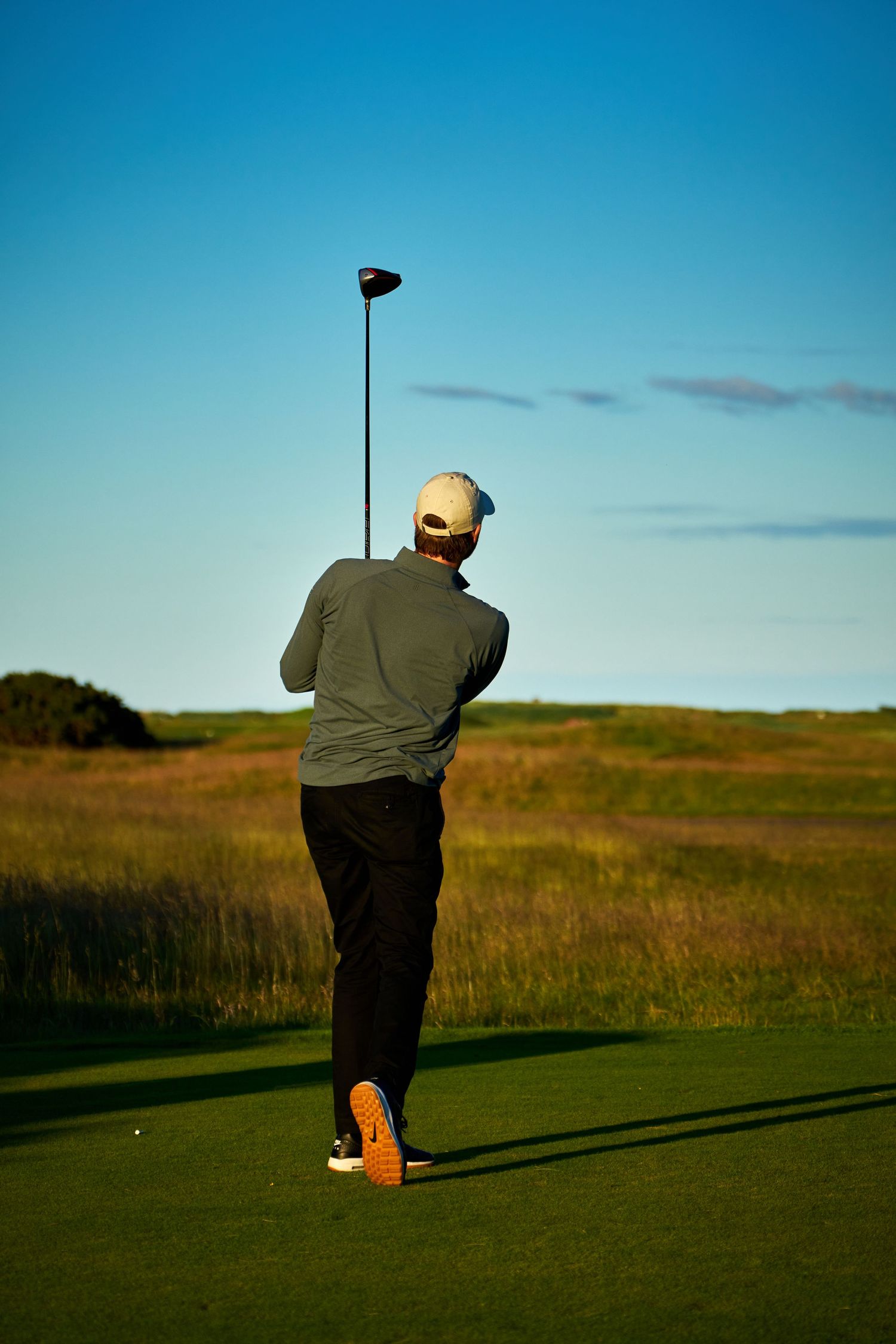 Golfer completing a follow-through swing on a scenic links golf course under a clear blue sky.