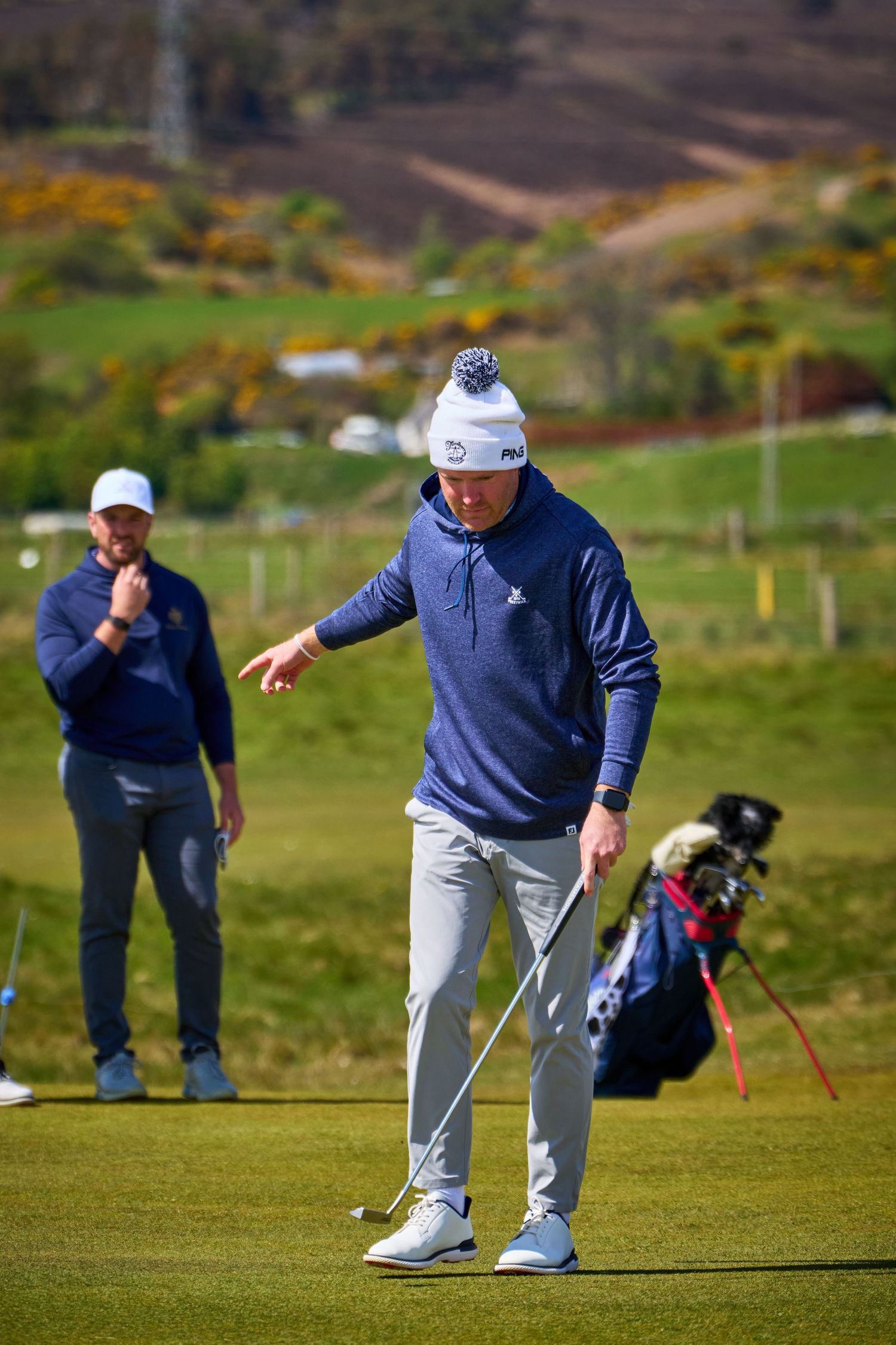 Golfer in navy FILA sweater and white beanie gestures on a golf course, with a caddie and golf bag in the background.