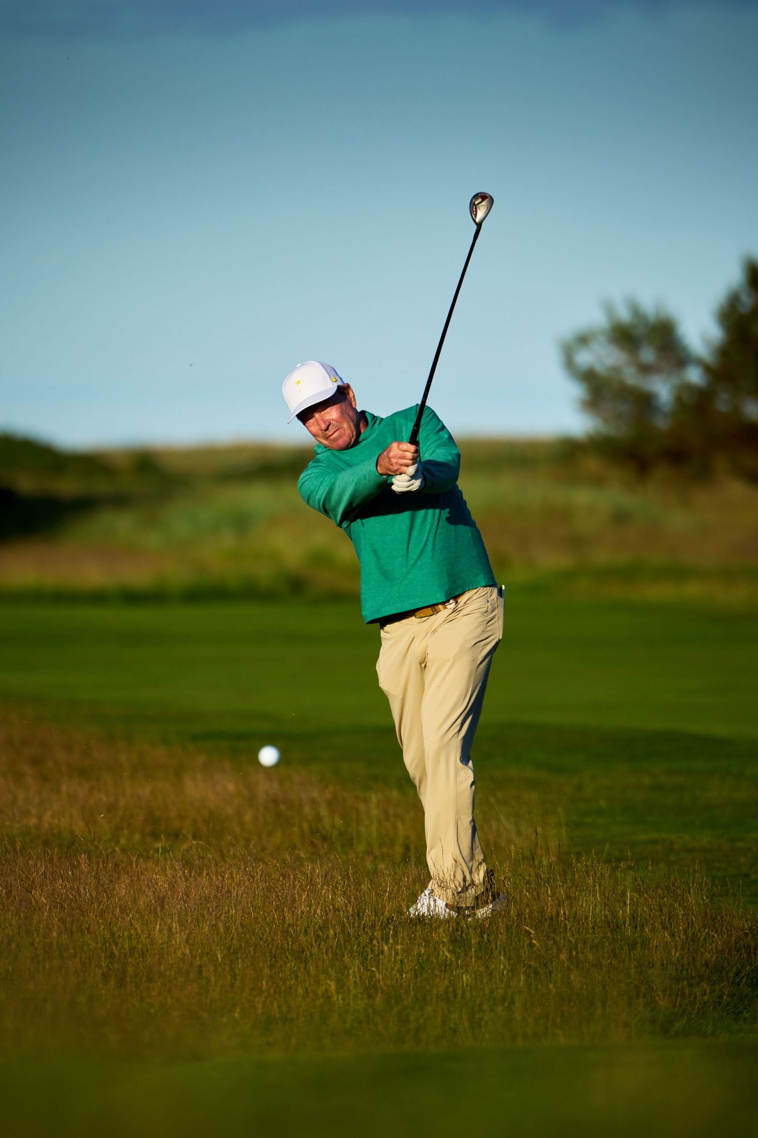 Golfer in green jacket and white cap swings a club on a lush golf course under a clear blue sky.