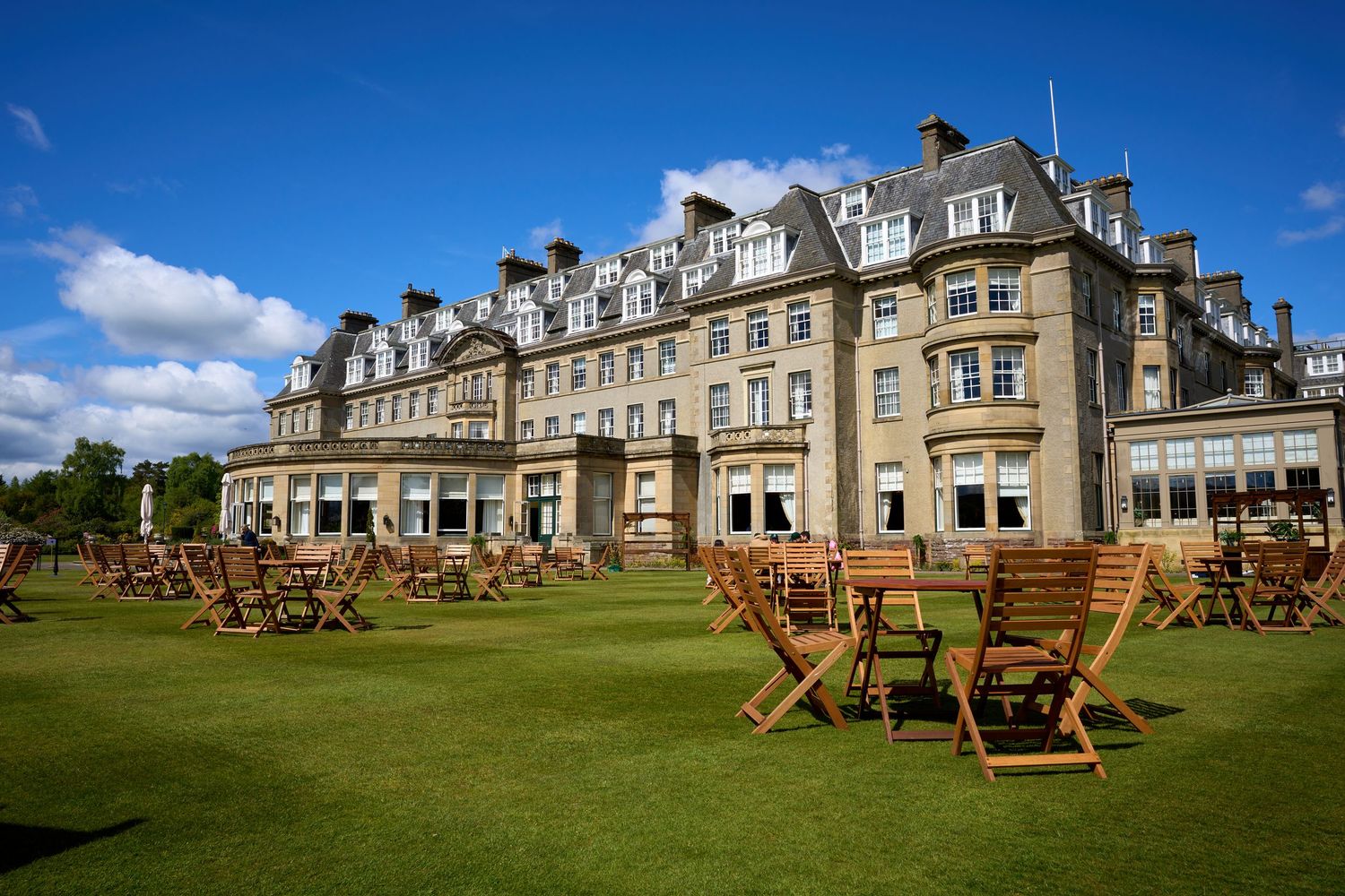 Elegant stone hotel with wooden outdoor furniture on lush green lawn under blue sky, Gleneagles Resort Scotland.