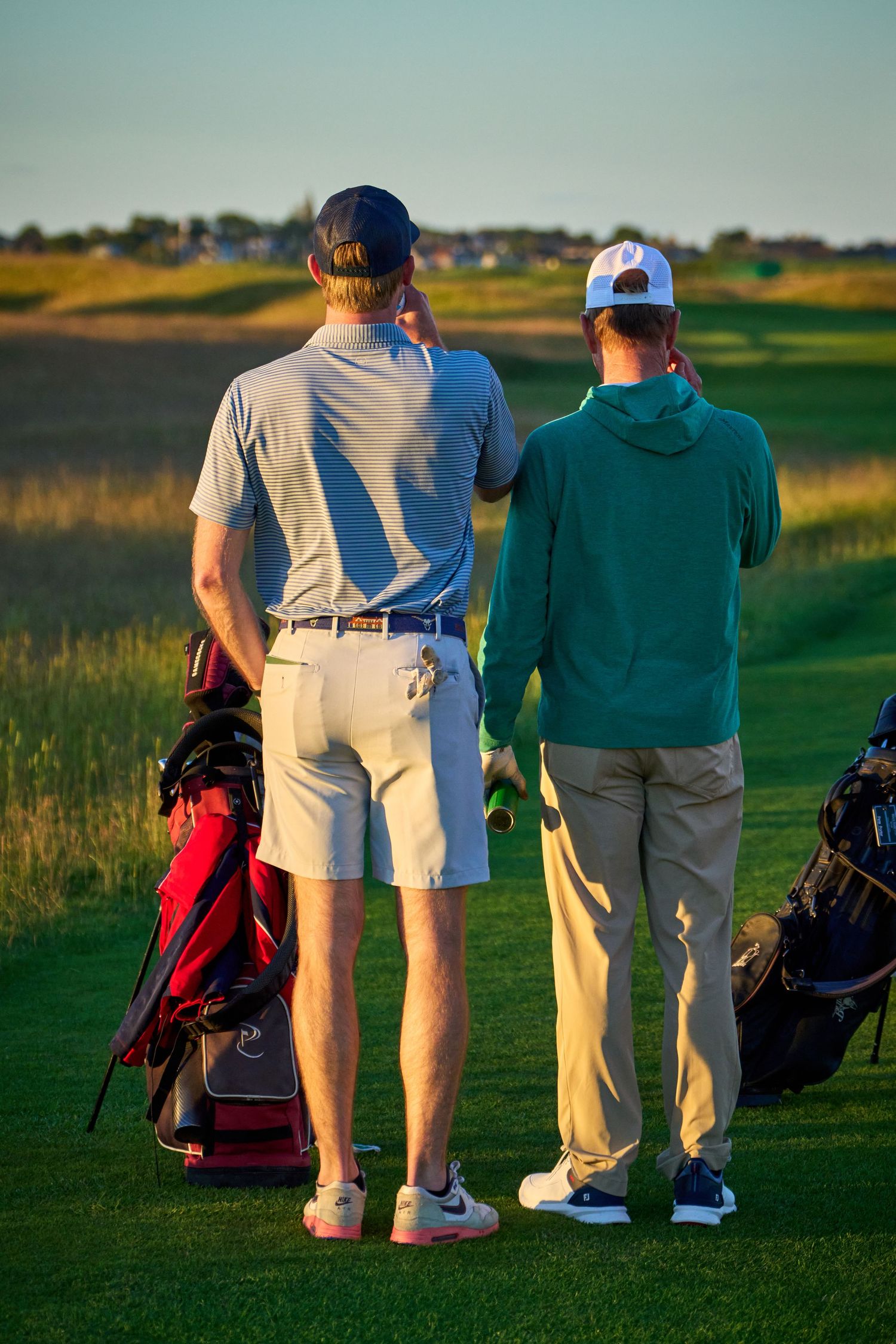 Two golfers stand on a sunny golf course reviewing their next shot, with golf bags nearby.
