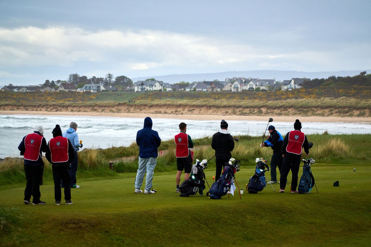 Golfers and caddies stand on a coastal links golf course fairway with sandy beach and seaside village in background.