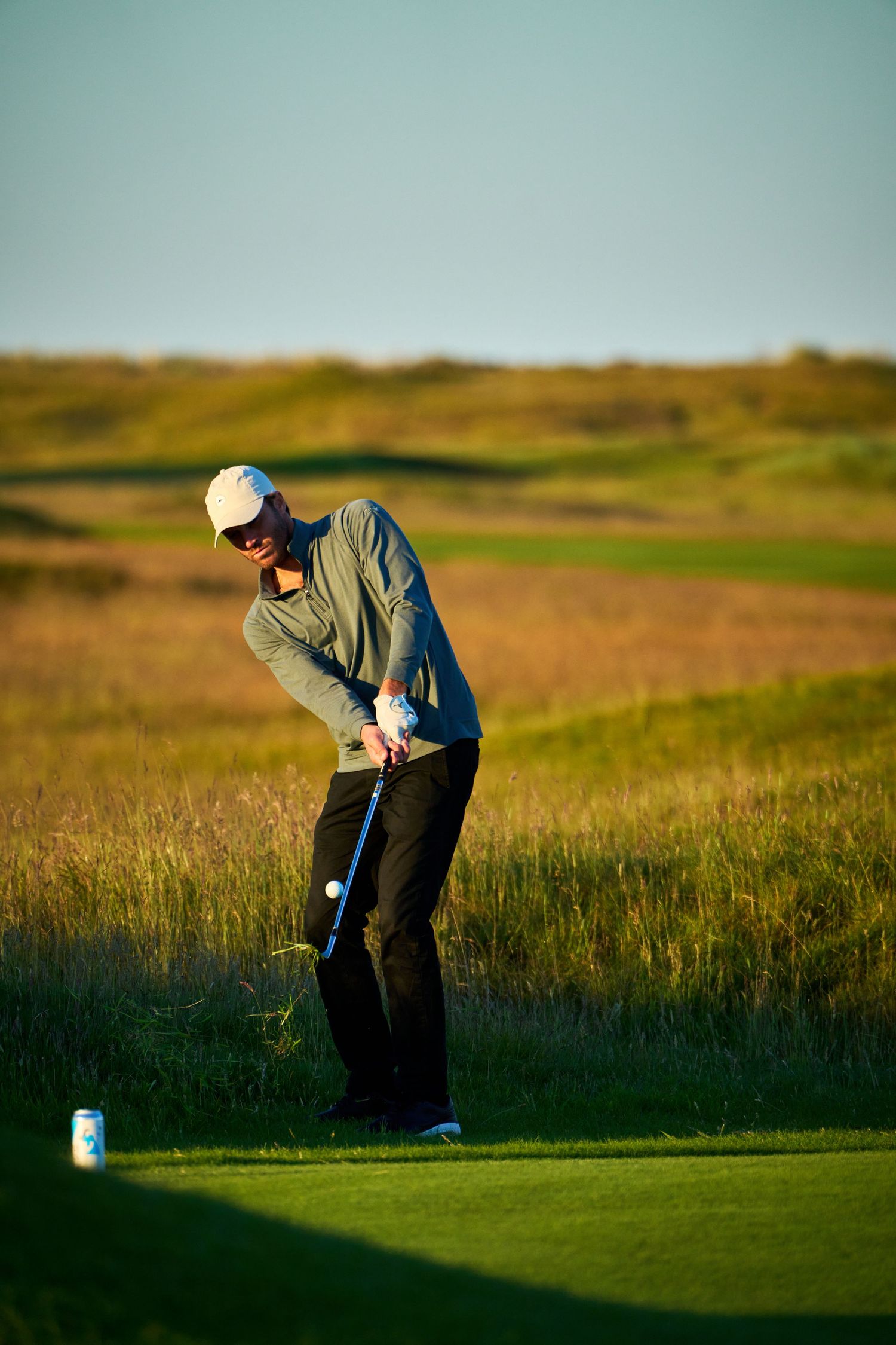 Golfer swinging a club on a scenic links golf course during golden hour with tall grass in the background.