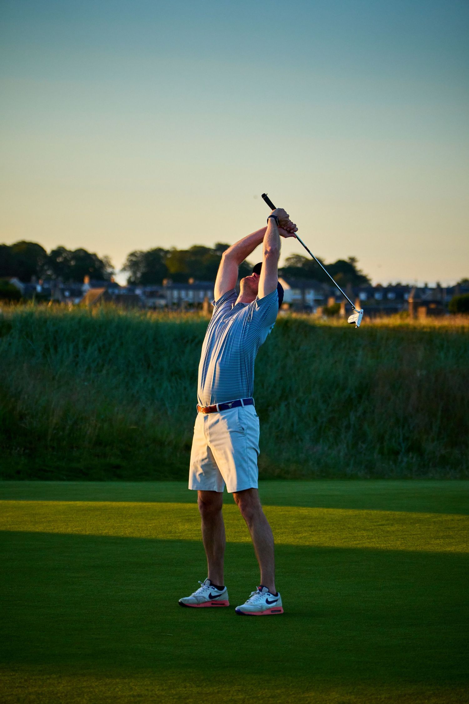Golfer completing a full swing on a lush green golf course at golden hour, wearing white shorts and Nike shoes.