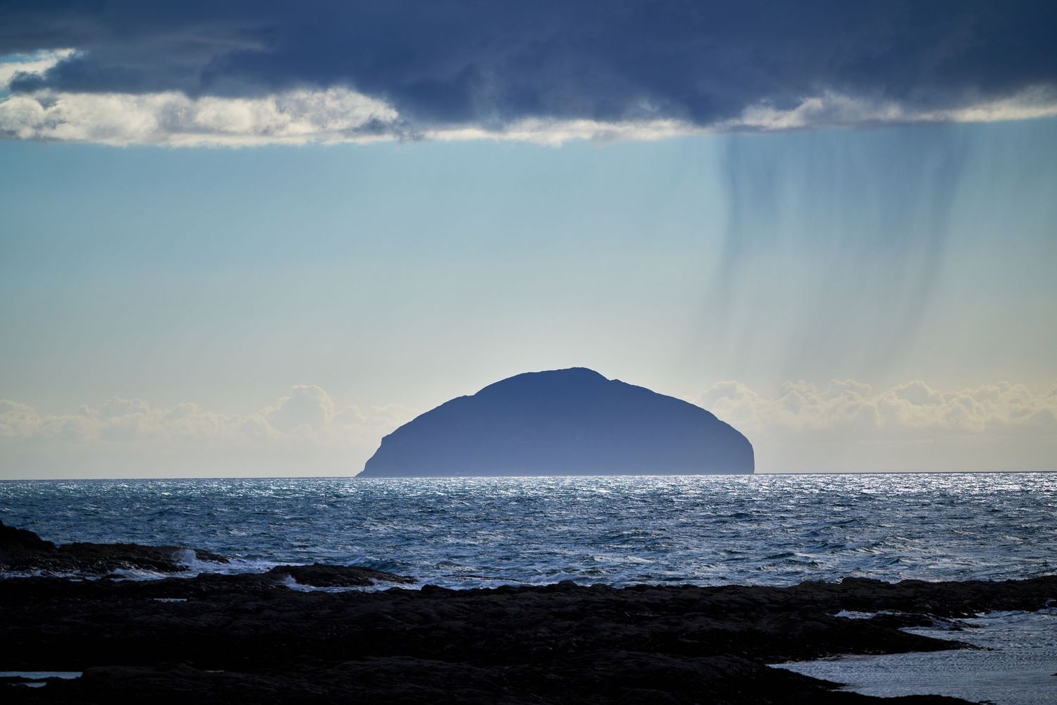 Dramatic silhouette of a small island rising from calm sea waters under stormy rays of light.