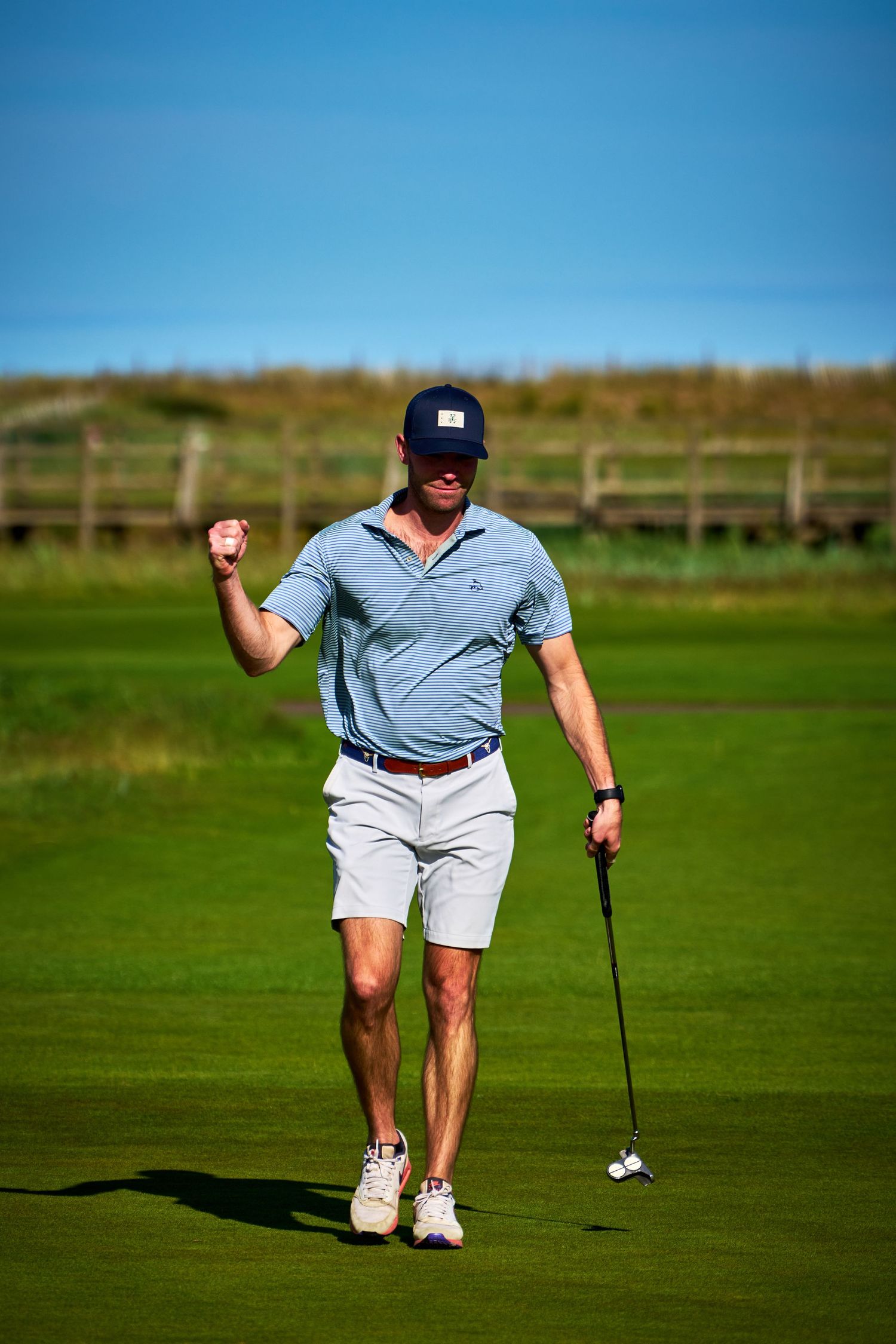 A golfer in a blue polo and cap pumps his fist in celebration while holding a putter on a sunny golf course.