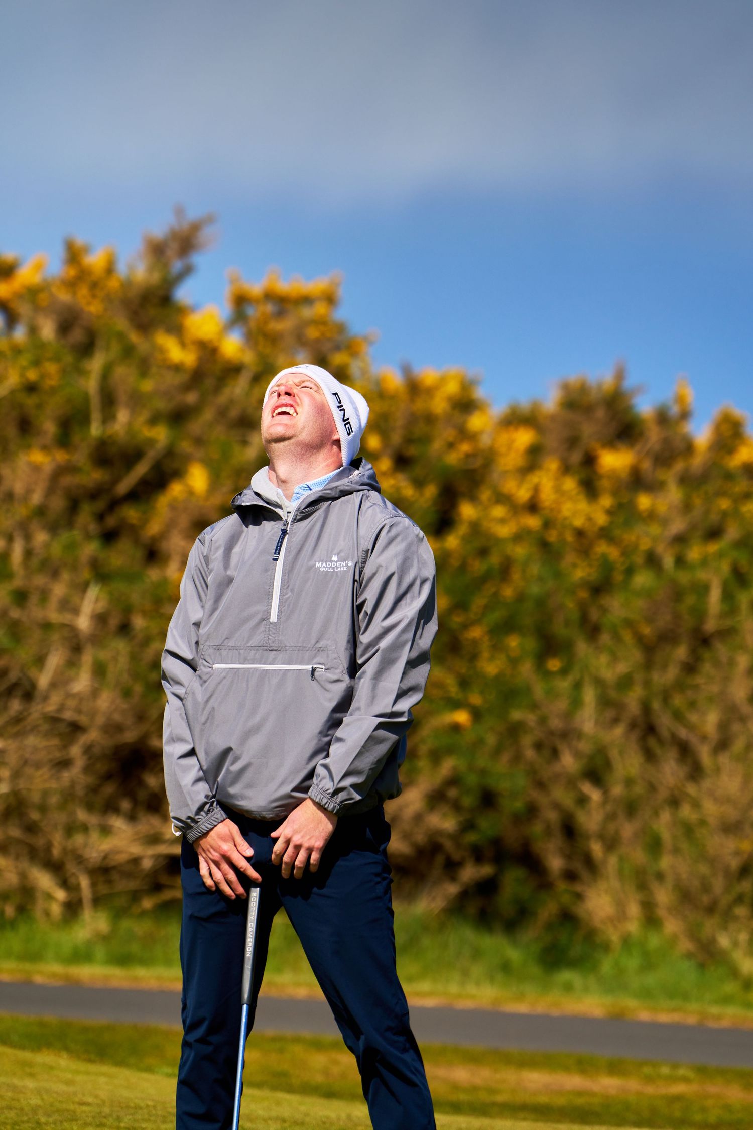 Golfer in gray jacket and white beanie laughs on golf course with yellow gorse bushes and blue sky.