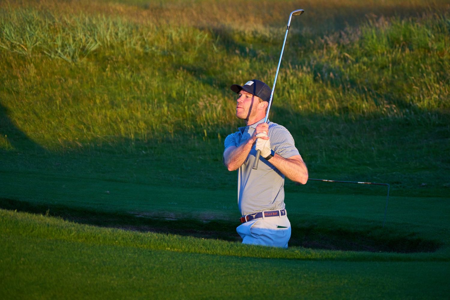 Golfer hitting a shot from a sand bunker on a golf course with green grass and golden light.
