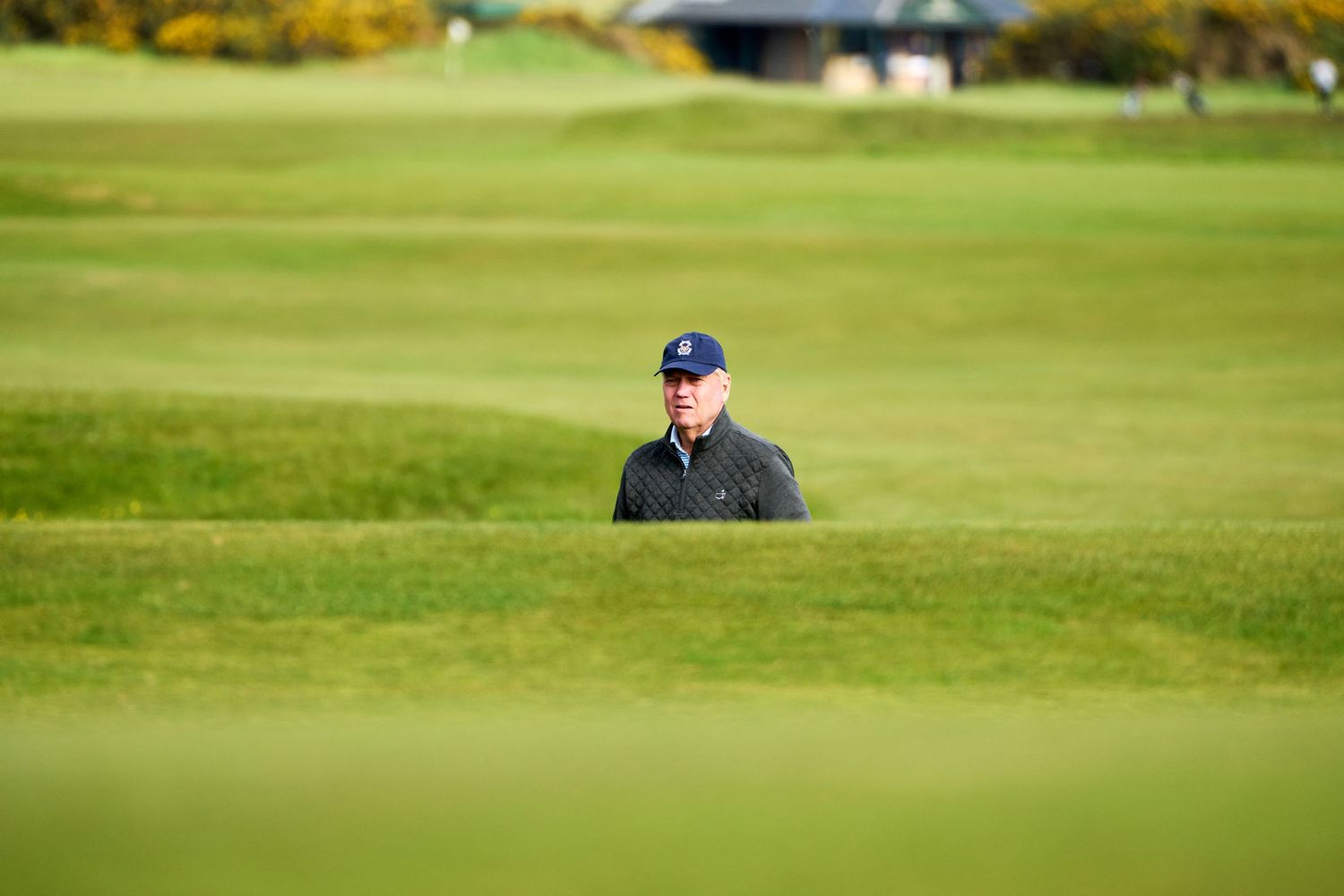 A golfer in a dark jacket and cap stands on a lush green golf course fairway on a cloudy day.