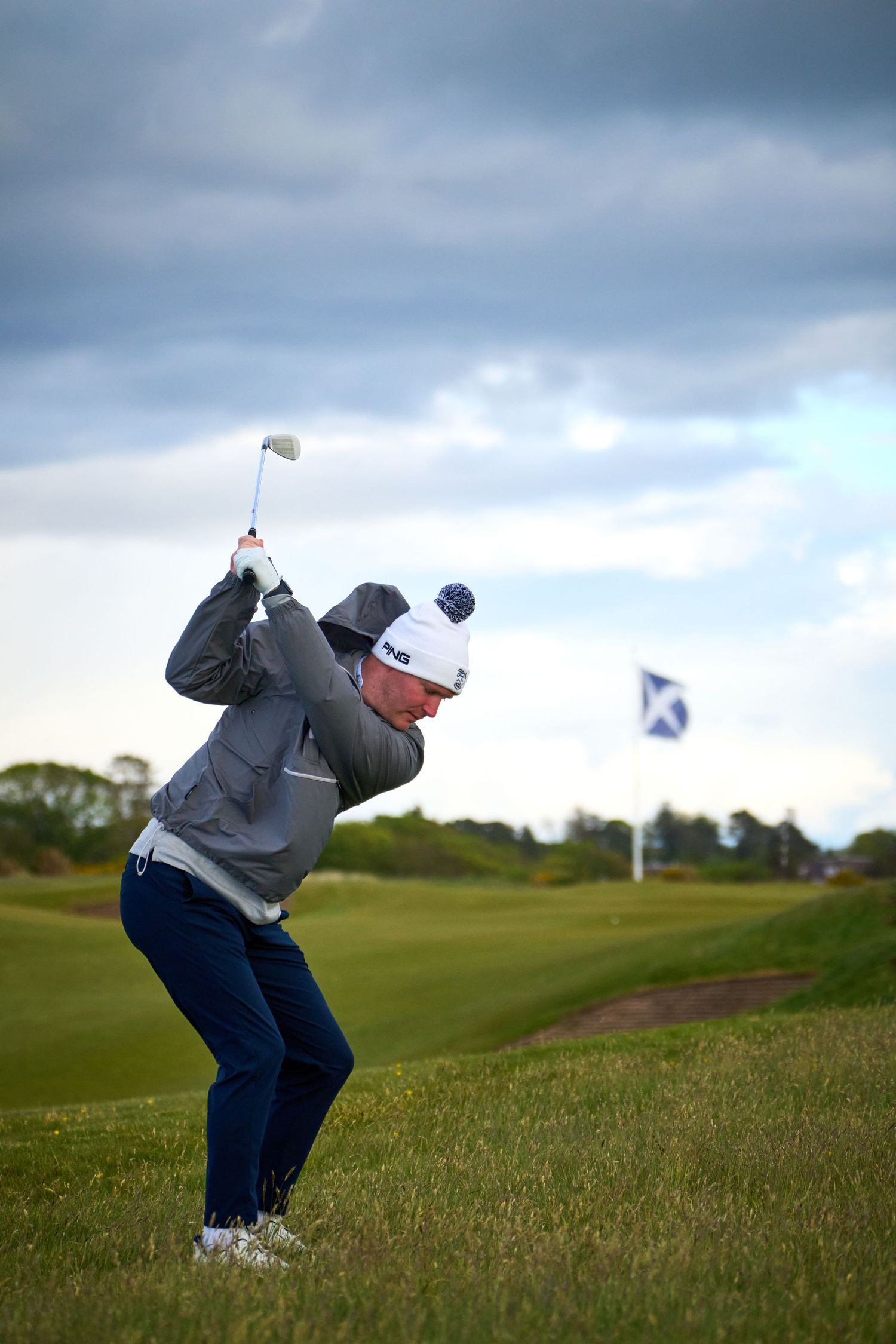 Golfer mid-swing on a lush green course with a Scottish flag in the background under cloudy skies.