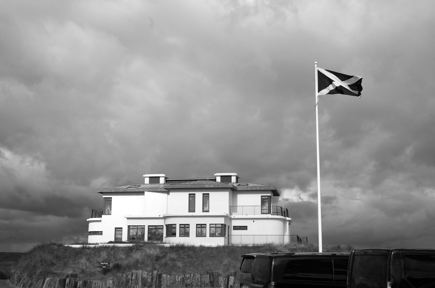 Black and white photo of a white building on a cliff with a Scottish Saltire flag flying against a dramatic cloudy sky.