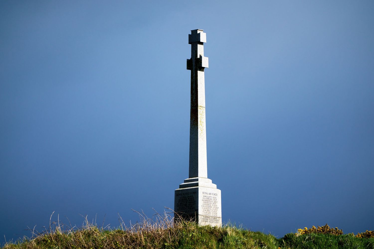 Stone cross monument atop a grassy hill against a dramatic deep blue sky.
