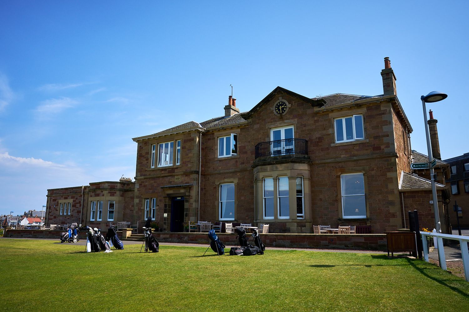 Historic red sandstone golf clubhouse with manicured green lawn under blue sky, likely St Andrews Links in Scotland.