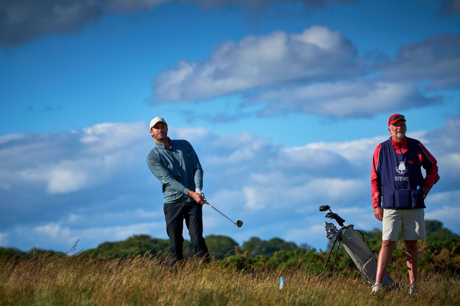 Two golfers on a links course, one mid-swing with a club, caddie standing beside a golf bag under a cloudy sky.
