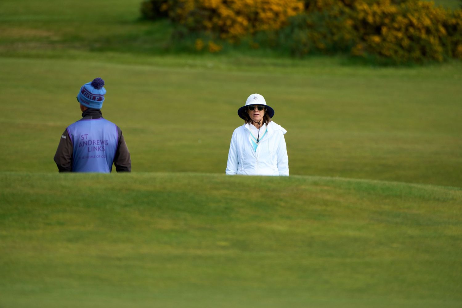 Two golfers standing on a lush green golf course, one wearing a blue hat and vest, the other in a white jacket and sun hat.