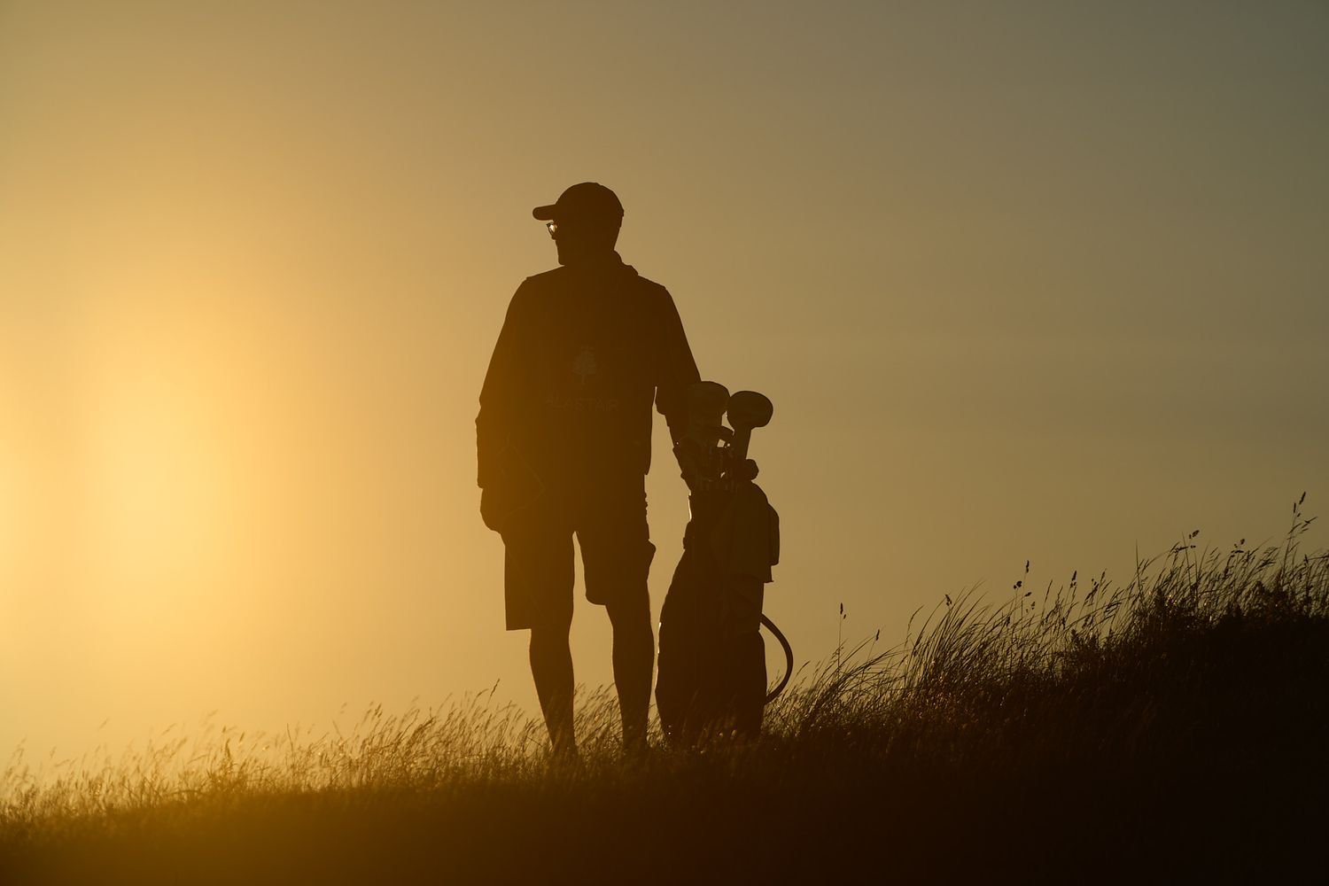 Silhouette of a golfer standing with a golf bag on a misty hill at sunset with golden sky.