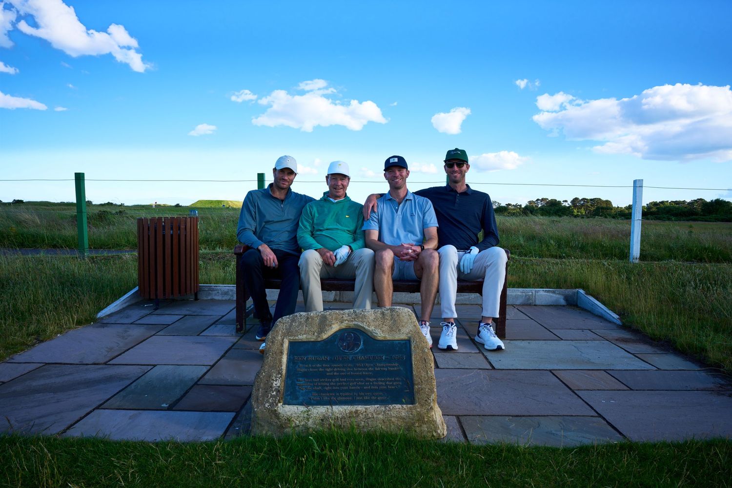 Four golfers pose on a stone bench beside a commemorative plaque on a scenic golf course under a blue sky.