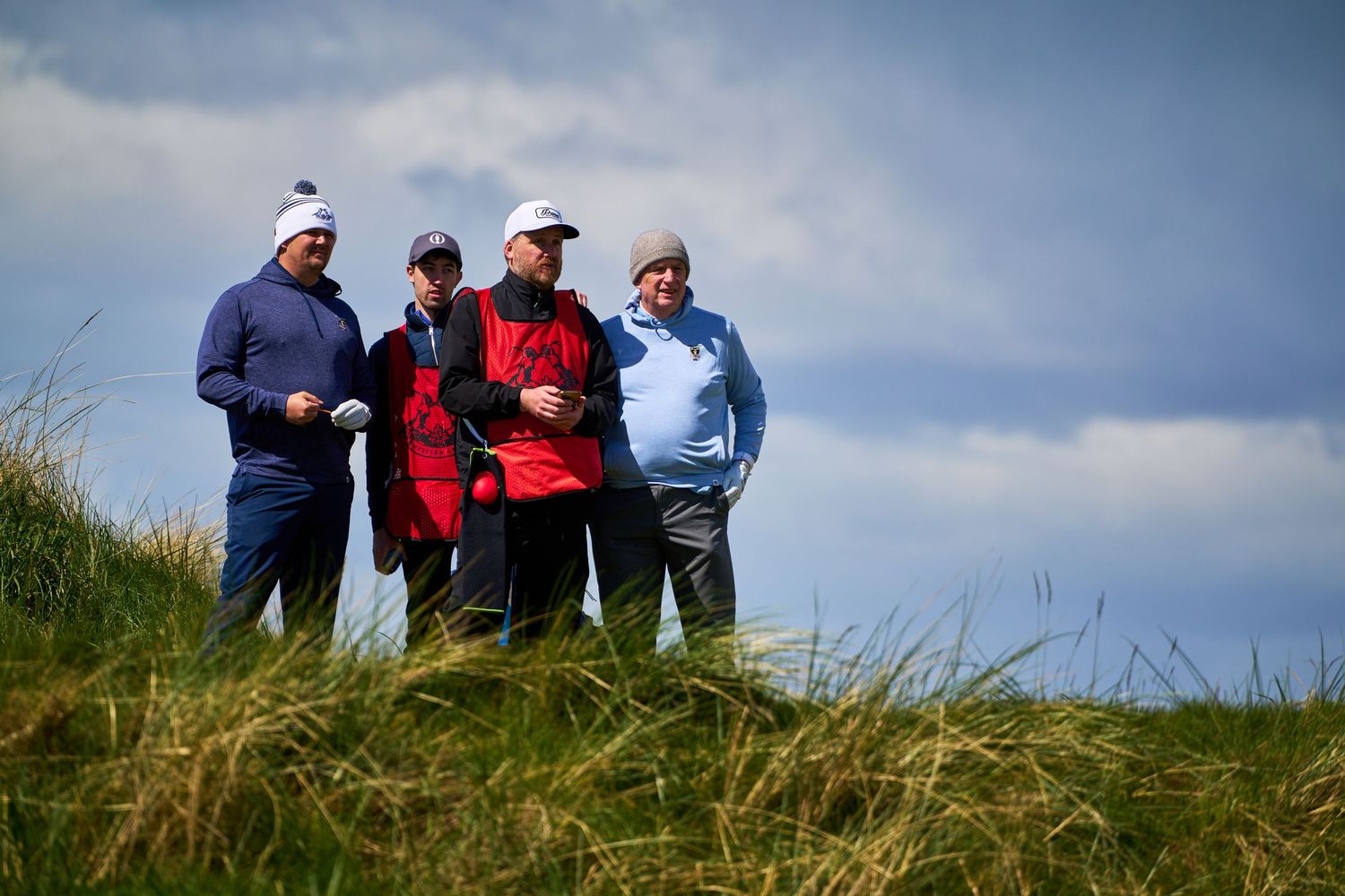 Four golfers standing on a grassy dune discussing strategy under a cloudy sky on a links golf course.