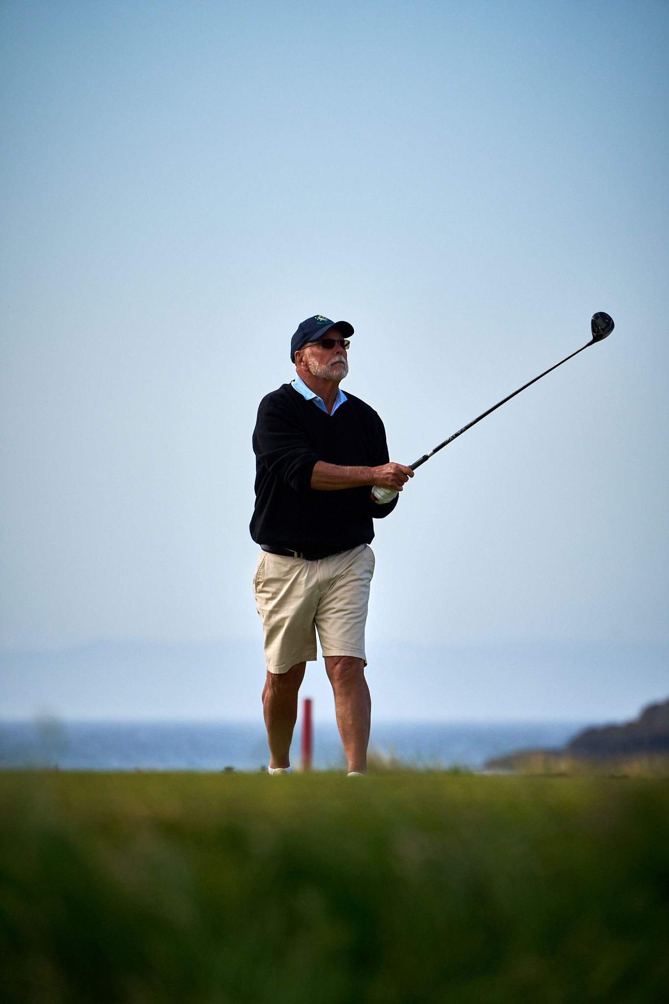 Senior male golfer completing a swing on a coastal golf course, wearing a navy sweater, khaki shorts, and cap.