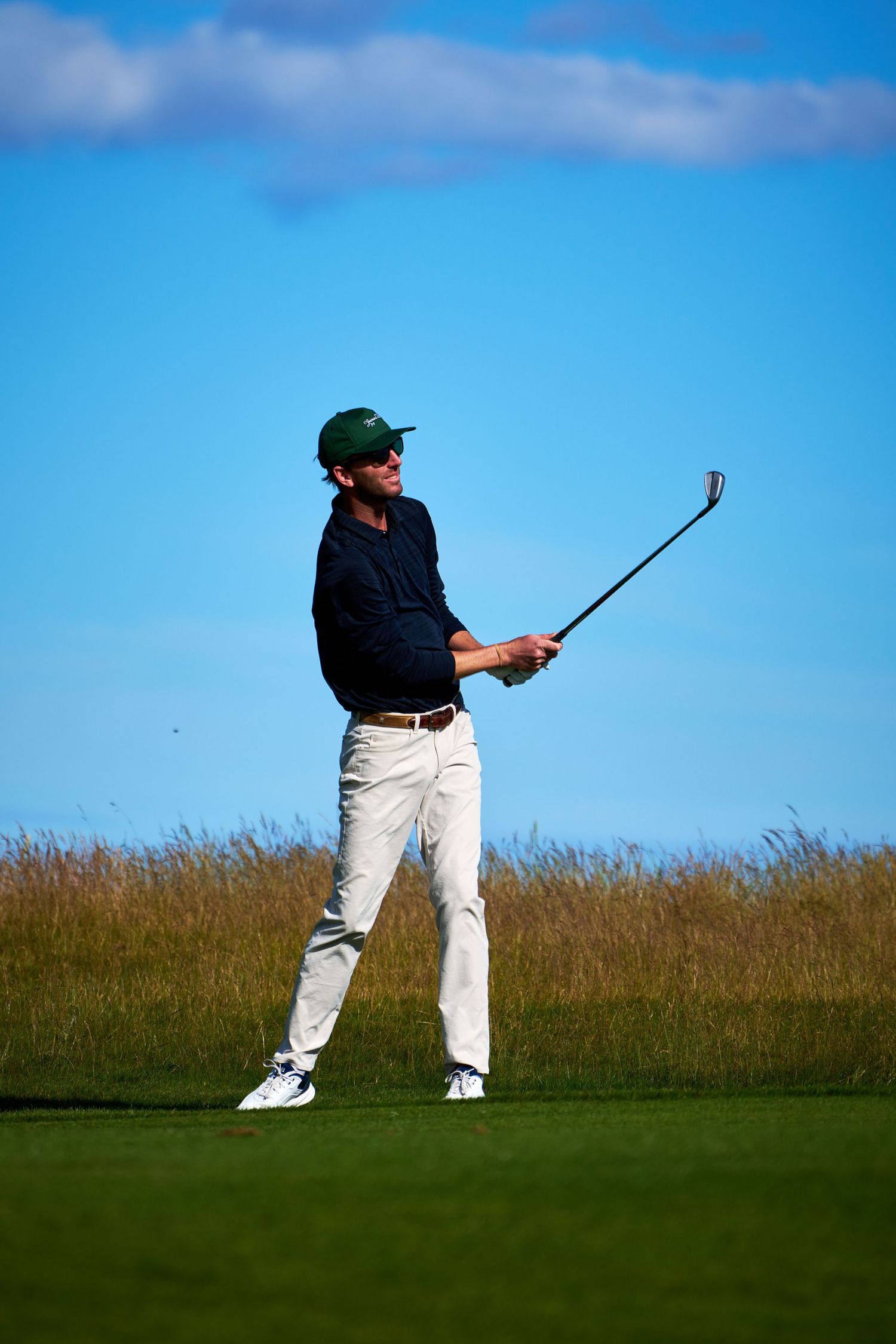Golfer in white pants and green cap follows through on an iron shot on a sunny golf course.