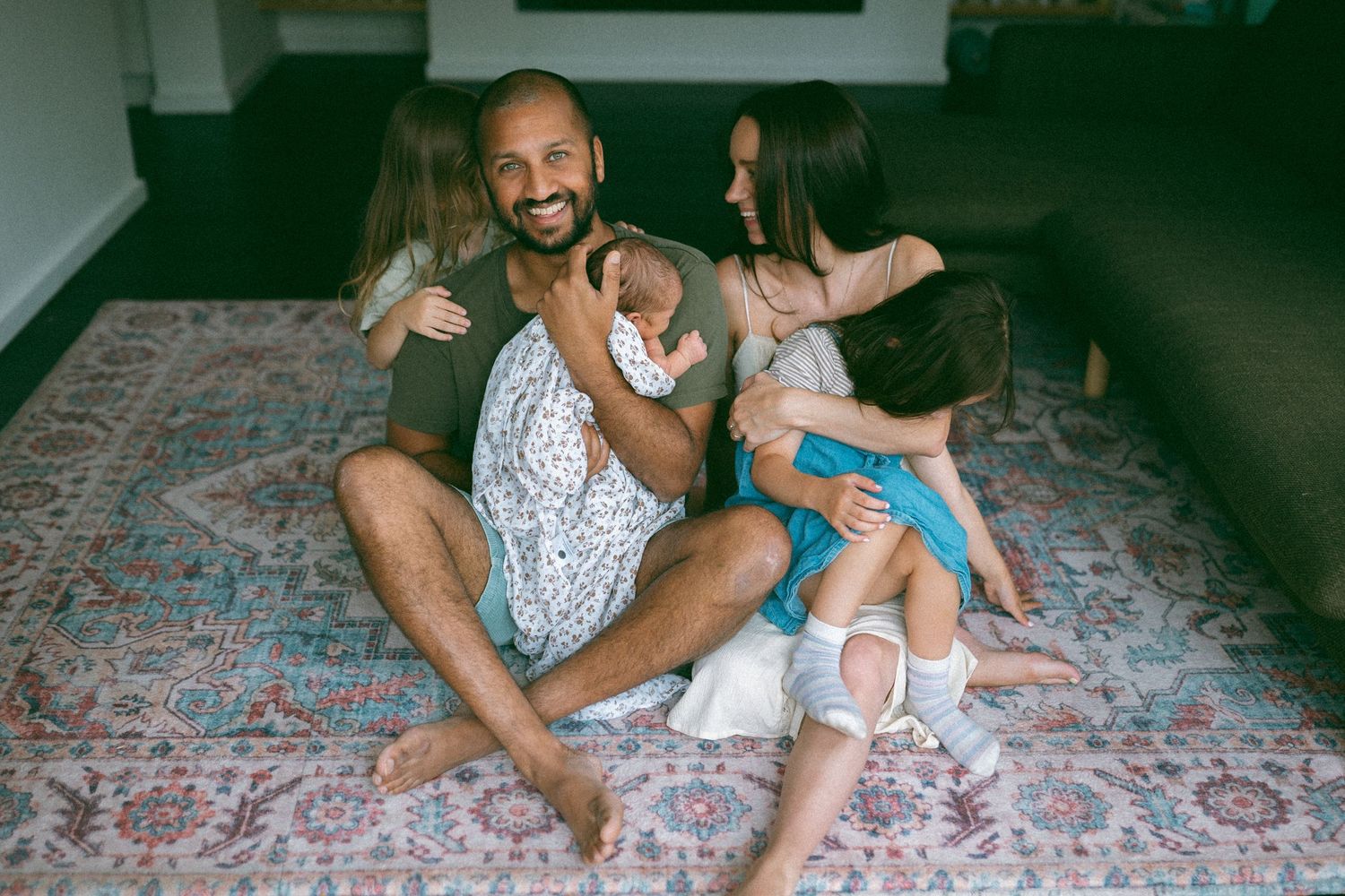 Happy family sitting on a rug, father smiling with children and mother cuddling together in a cozy living room.