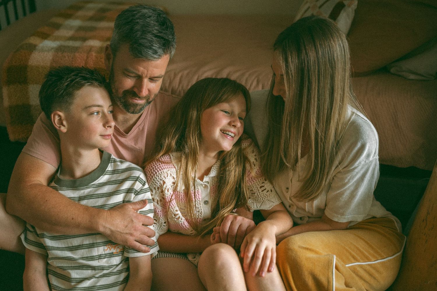 A happy family of four sitting together on the floor, laughing and embracing in a warmly lit bedroom.