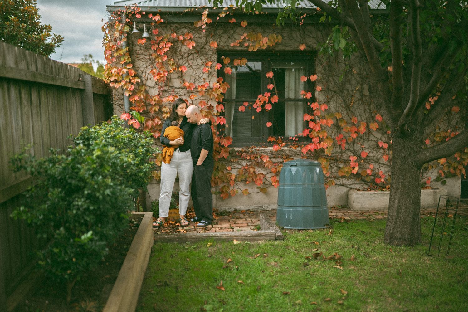 Couple embracing in autumn garden beside ivy-covered cottage with colorful fall foliage and green lawn.
