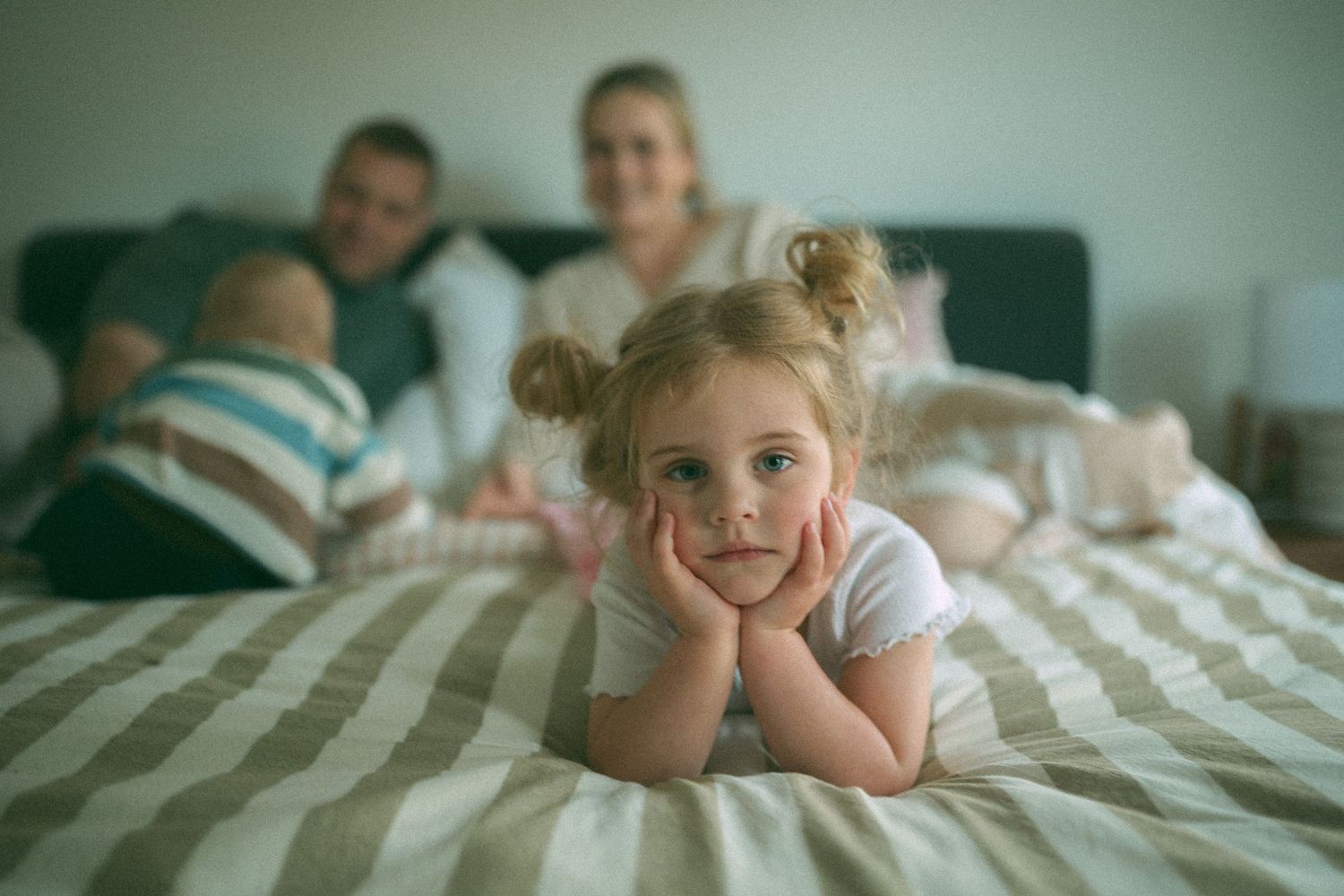 Young girl resting chin on hands on bed, smiling, with parents and baby sibling relaxing in background.
