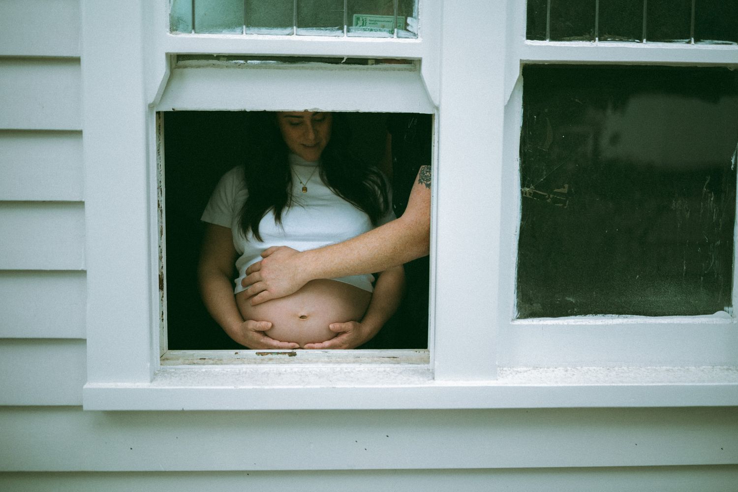 Pregnant woman standing at an open window, cradling her baby bump with both hands, wearing a white t-shirt.