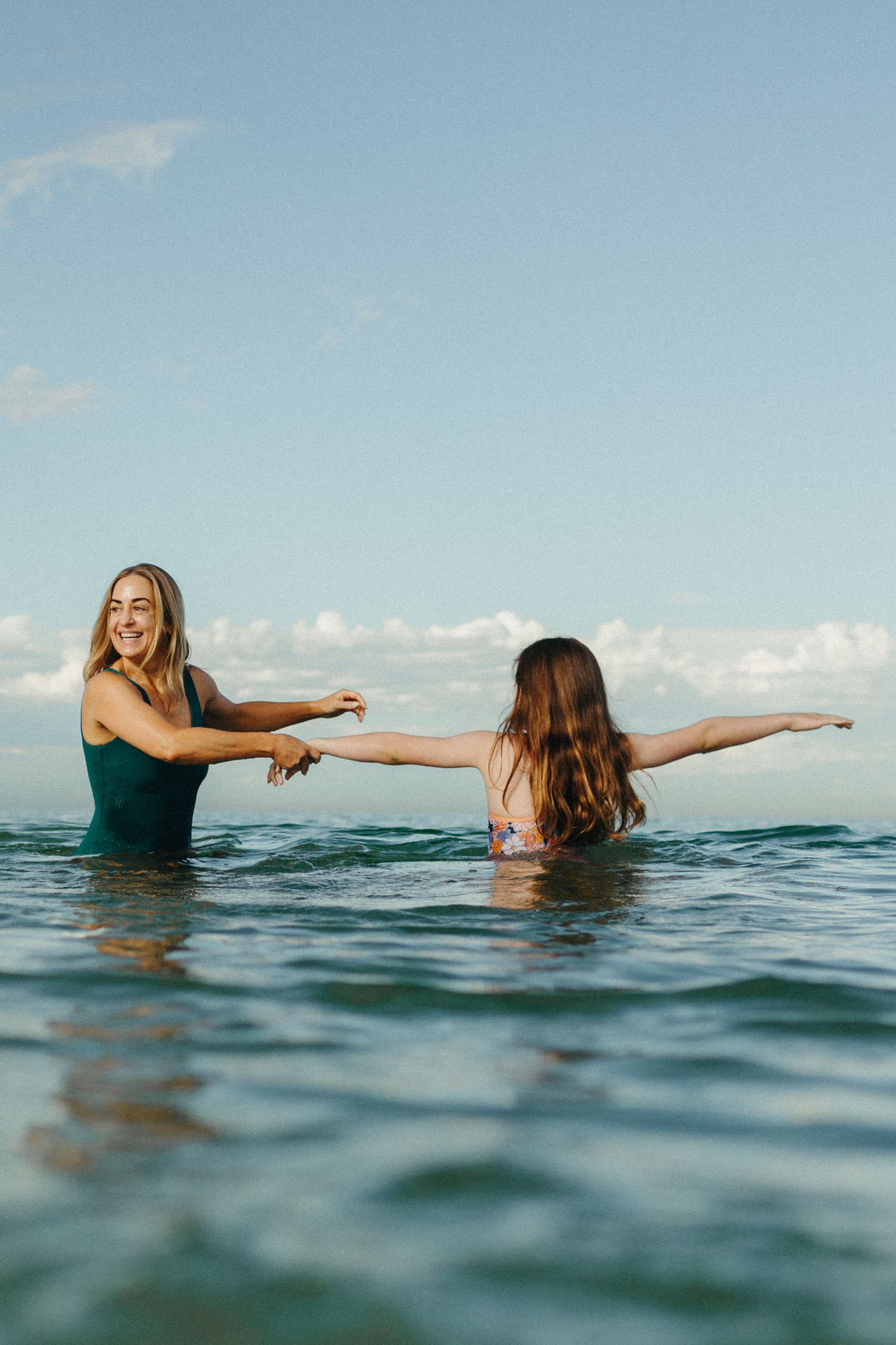 Two women enjoying the ocean, one smiling in a green swimsuit holding hands with another in a pink bikini.