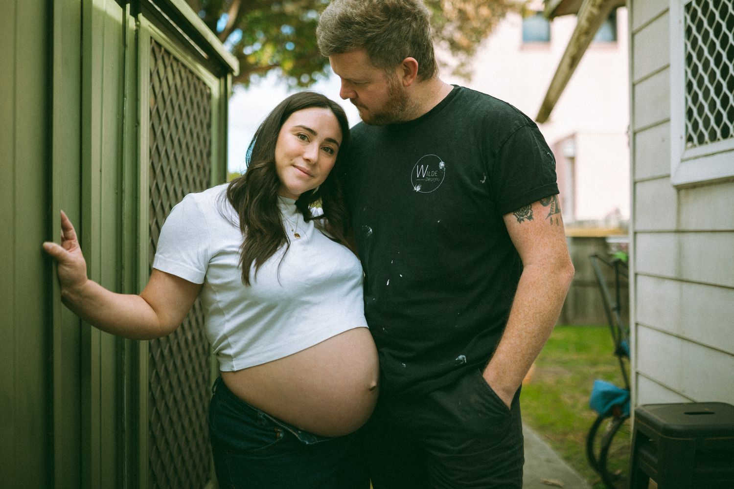 Pregnant couple sharing a tender moment outdoors, woman in white crop top showing baby bump, man in black t-shirt.