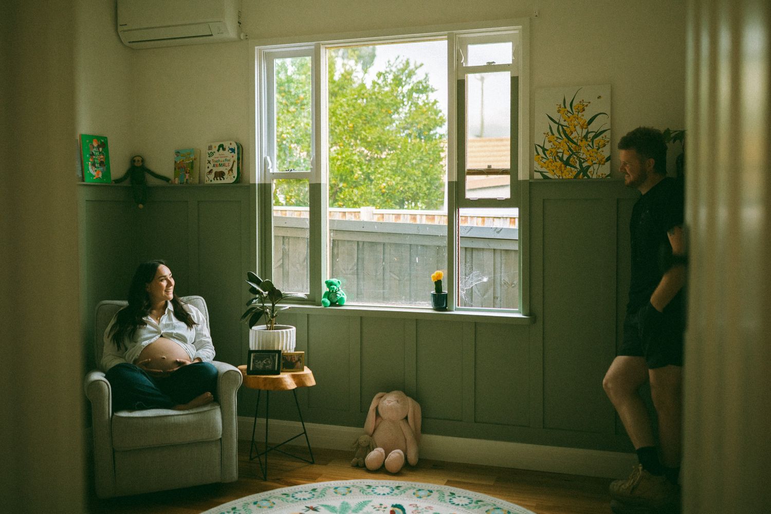 Pregnant woman relaxing in nursery chair while partner stands nearby in a cozy, decorated room with natural light.