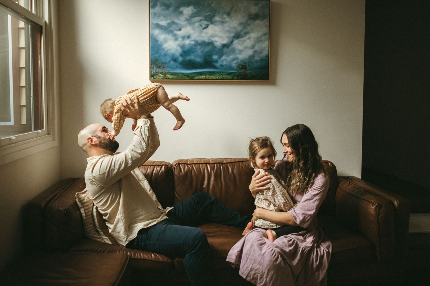 Family bonding on brown leather sofa, father lifting baby while mother holds toddler in cozy living room.