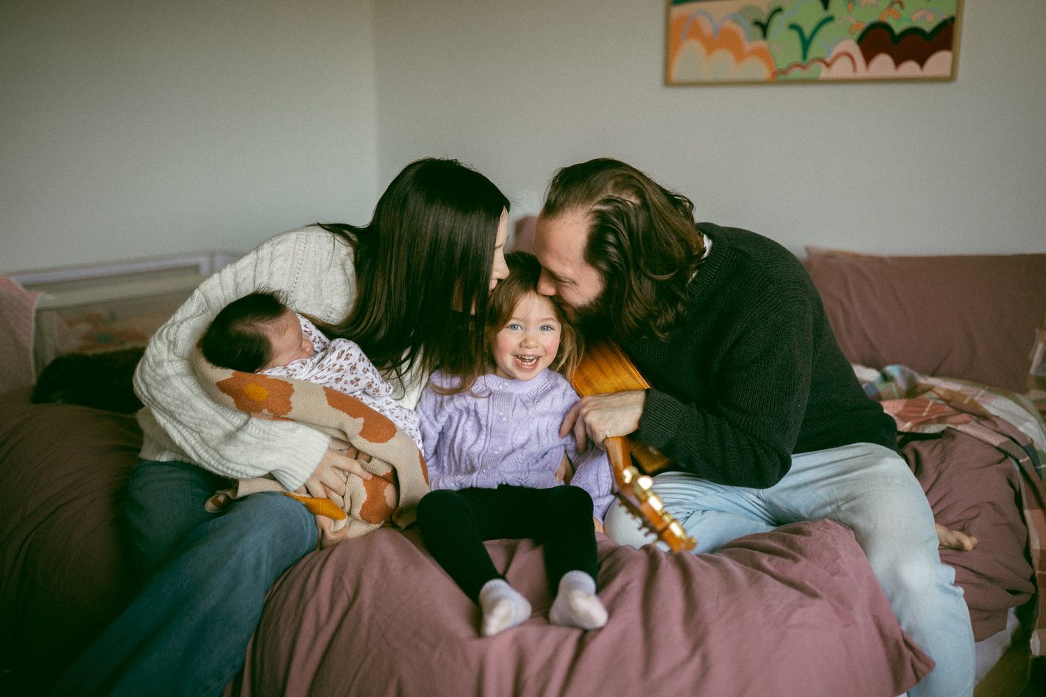 Family of four sitting on a couch, parents kissing toddler girl while holding a newborn baby at home.