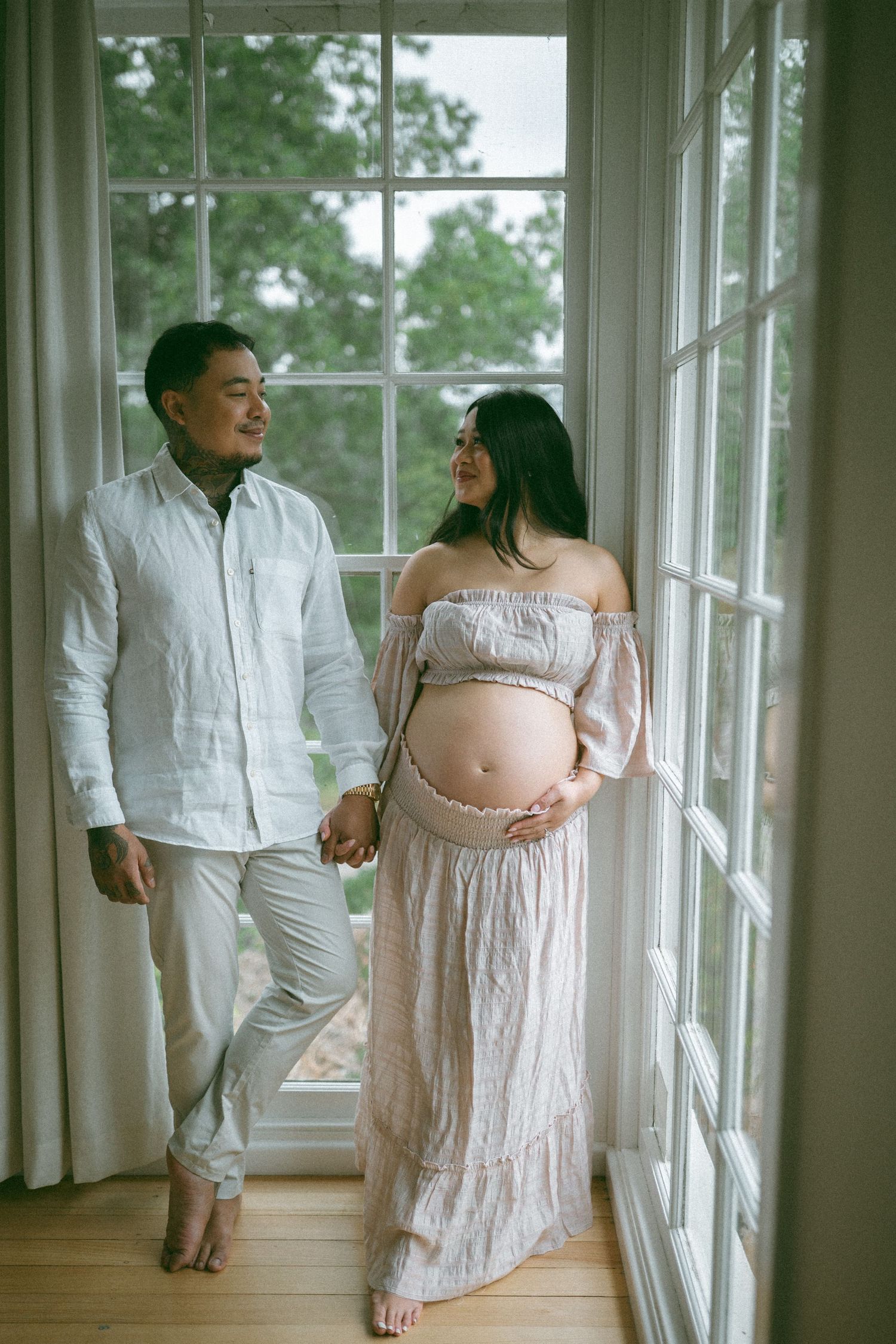 A couple poses by a large window, the pregnant woman in a flowy off-shoulder dress cradling her baby bump.