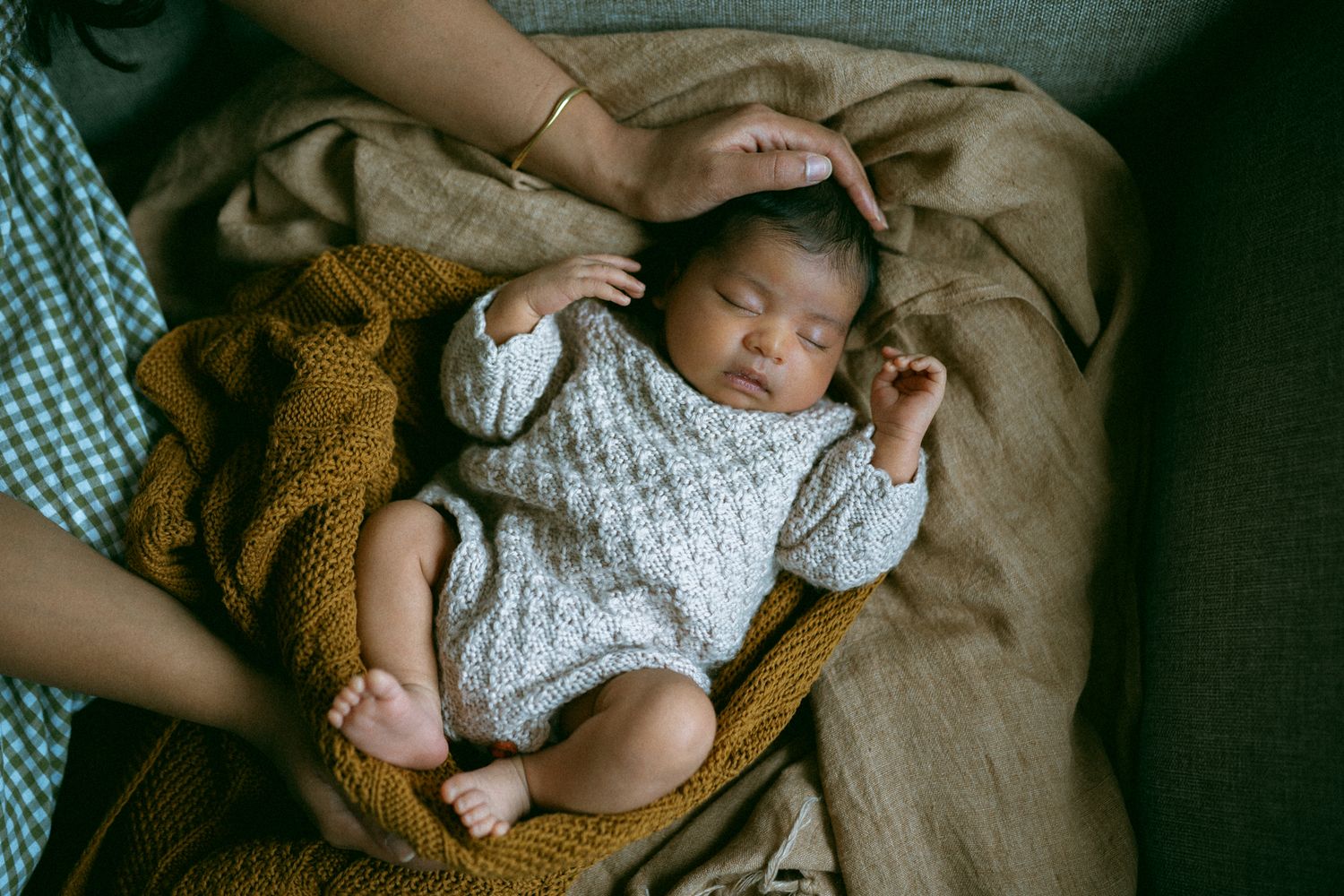 Sleeping newborn baby in a gray knit romper resting on a mustard yellow blanket, cradled by a parent's gentle hand.