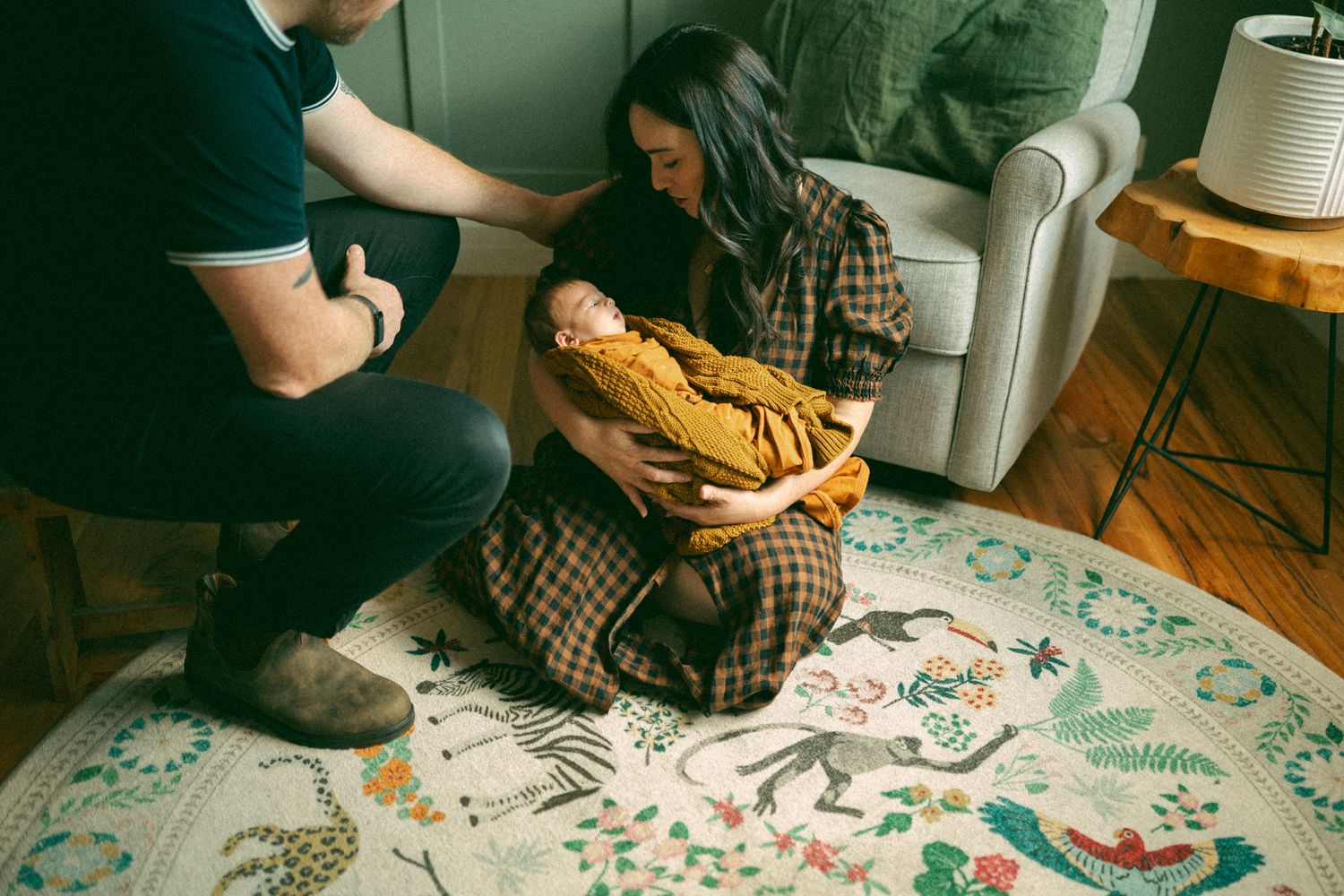 Parents sitting on a floral rug admiring their newborn baby wrapped in a mustard yellow swaddle blanket indoors.